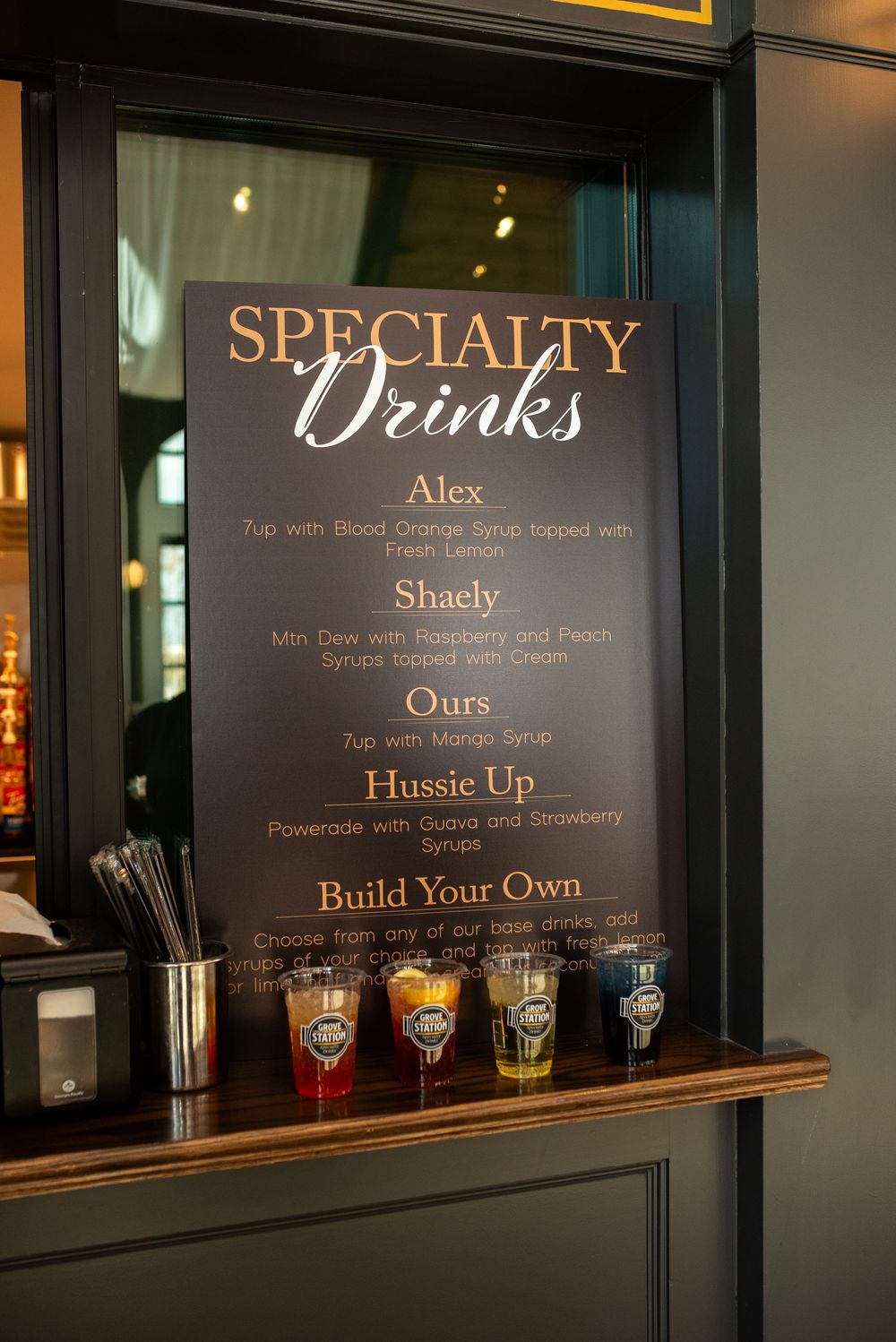 Illuminated drink menu board displaying cocktail names and options through a window at night.