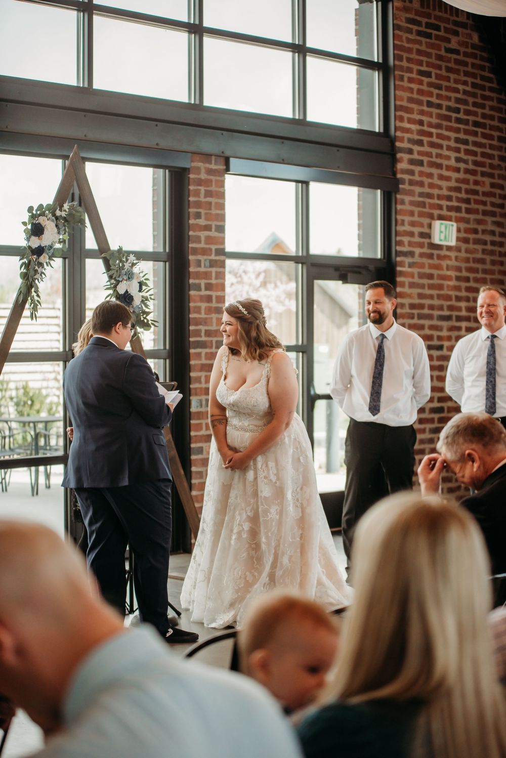 Wedding ceremony taking place in an industrial venue with brick walls and large windows as guests look on during the exchange.