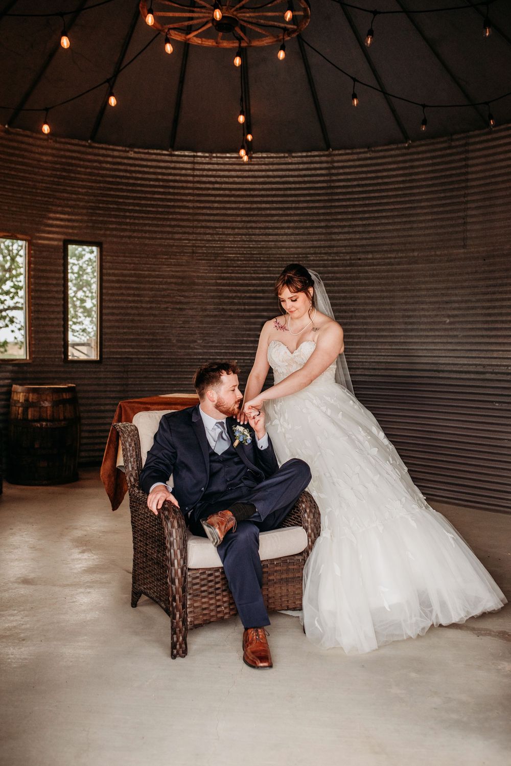Wedding couple sits together in a rustic barn venue with string lights creating a warm, romantic atmosphere.