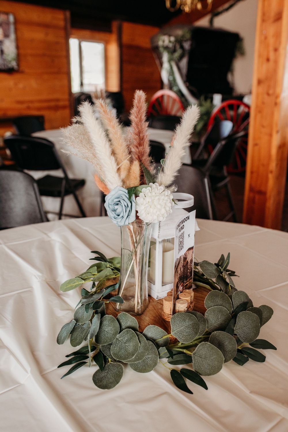Elegant table centerpieces featuring eucalyptus greenery, white pampas grass, and light blue floral accents on white tablecloths.