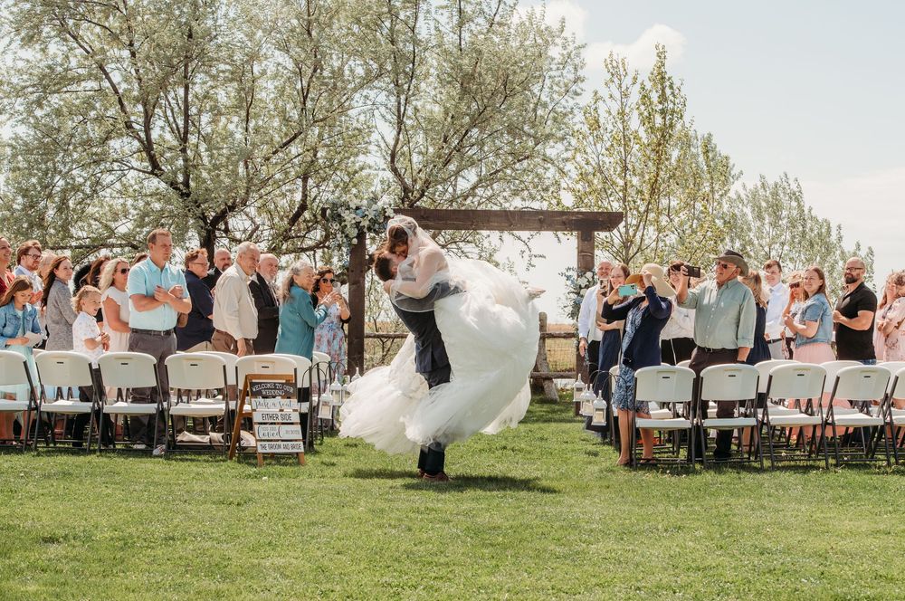 A couple performs a romantic dance lift during an outdoor wedding ceremony with guests seated on white chairs beneath trees.