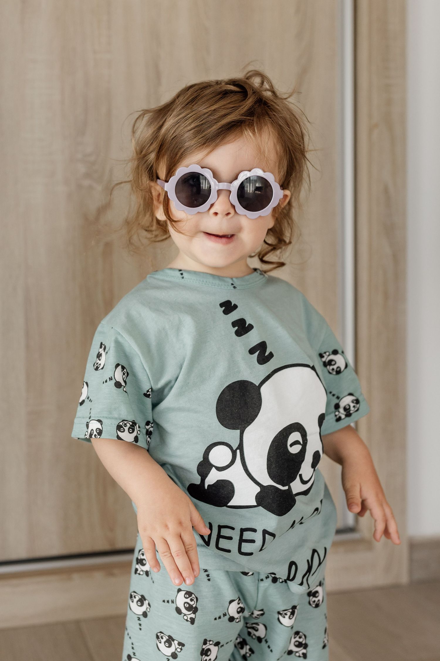 A toddler wearing round sunglasses and a mint green panda pajama set strikes a playful pose.