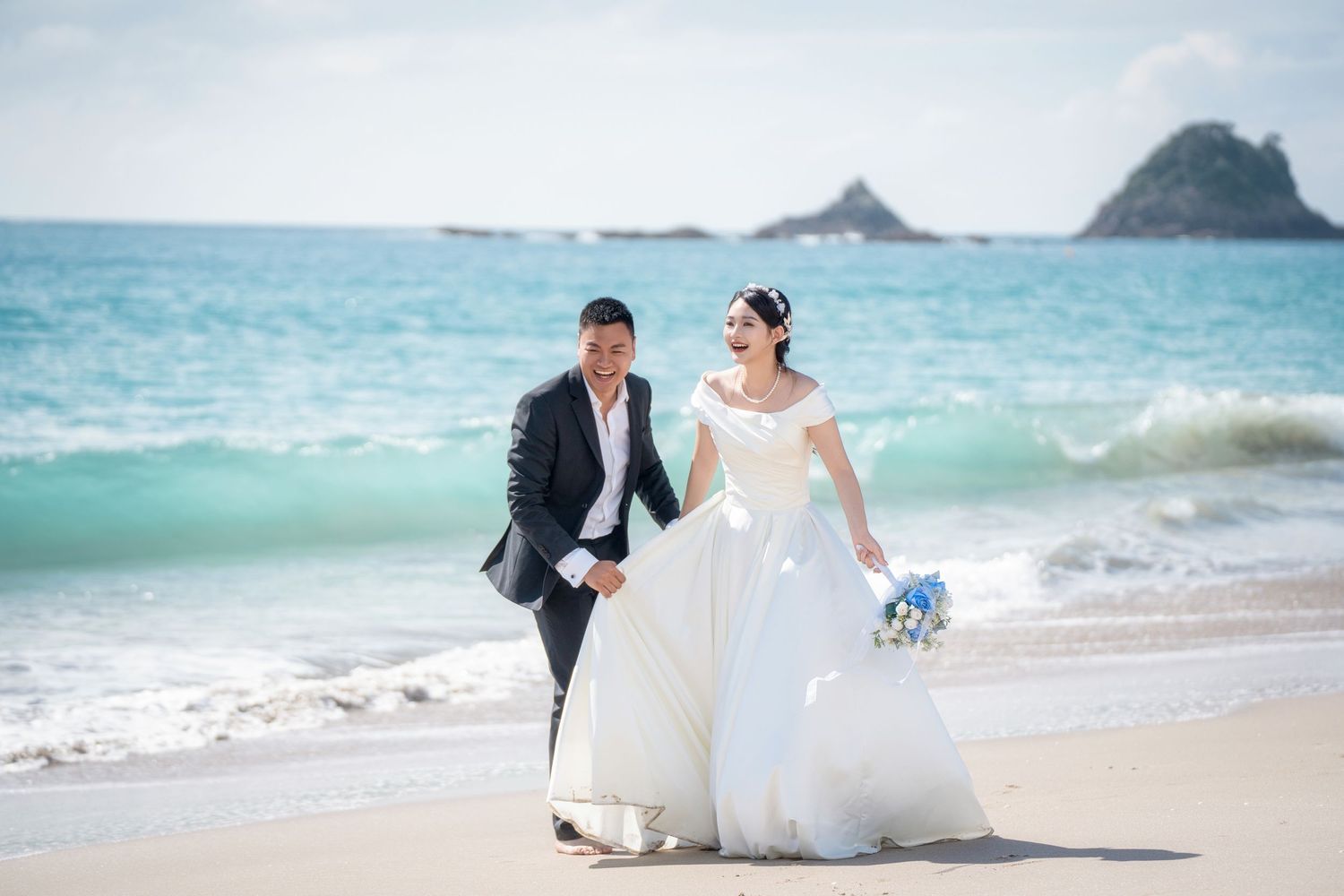 Happy bride and groom walking on a beautiful white sand beach with turquoise ocean waves for Auckland coastal wedding photography.
