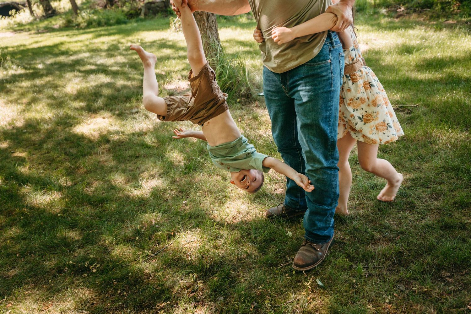 Adult playfully swing two young children upside down on a lush green lawn during a fun outdoor family activity.