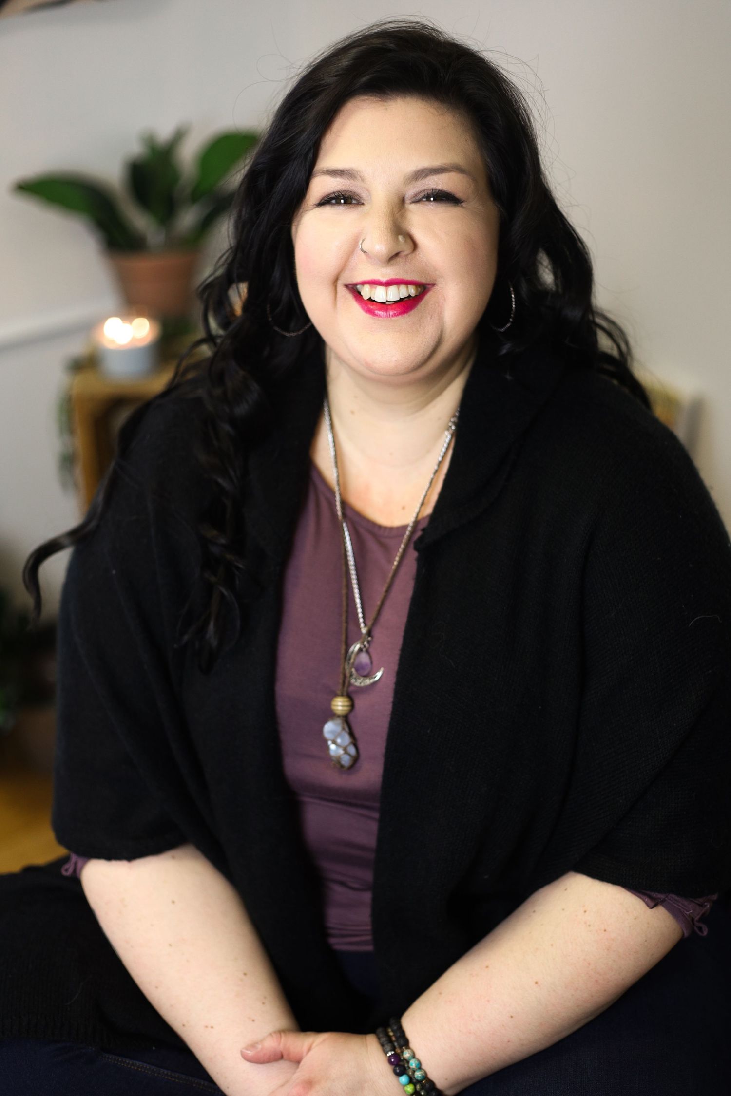 Professional portrait of a smiling person wearing a black cardigan and purple top with layered necklaces against a light background.