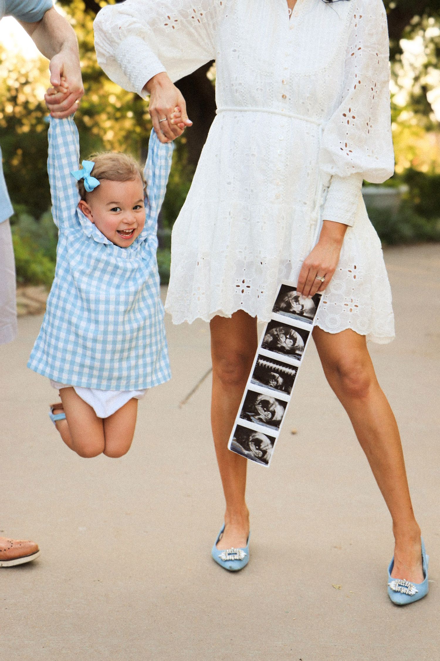 A small child in a blue gingham dress jumps joyfully while holding hands with someone in a white eyelet dress holding sonogram photos.