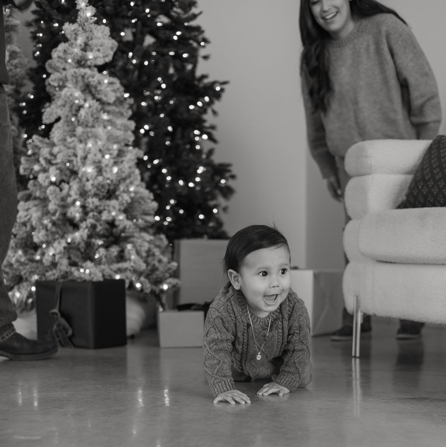 Baby crawling on floor near illuminated Christmas tree with parent watching from couch.