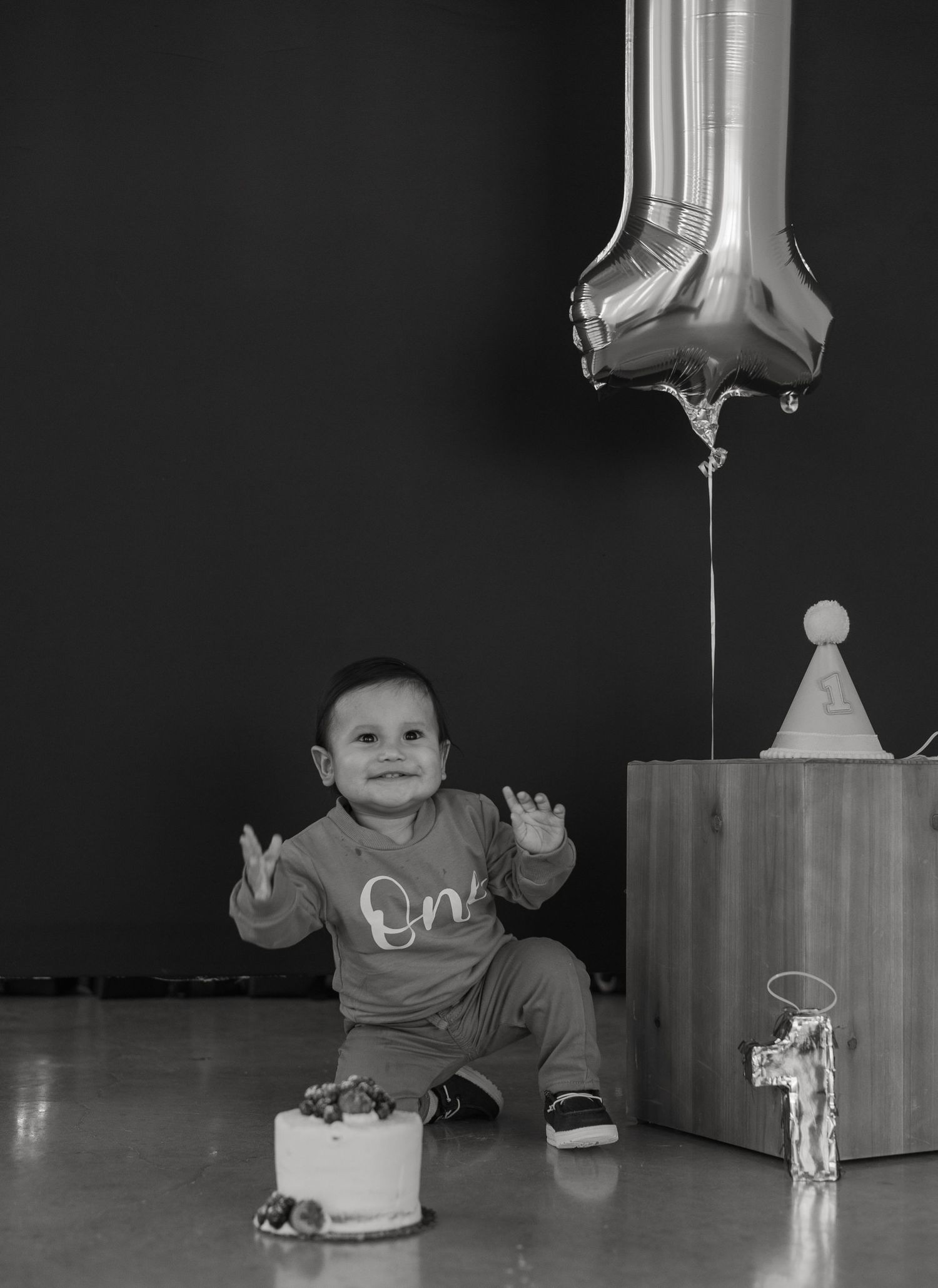 Baby celebrating first birthday with small cake and balloon in black and white photo.