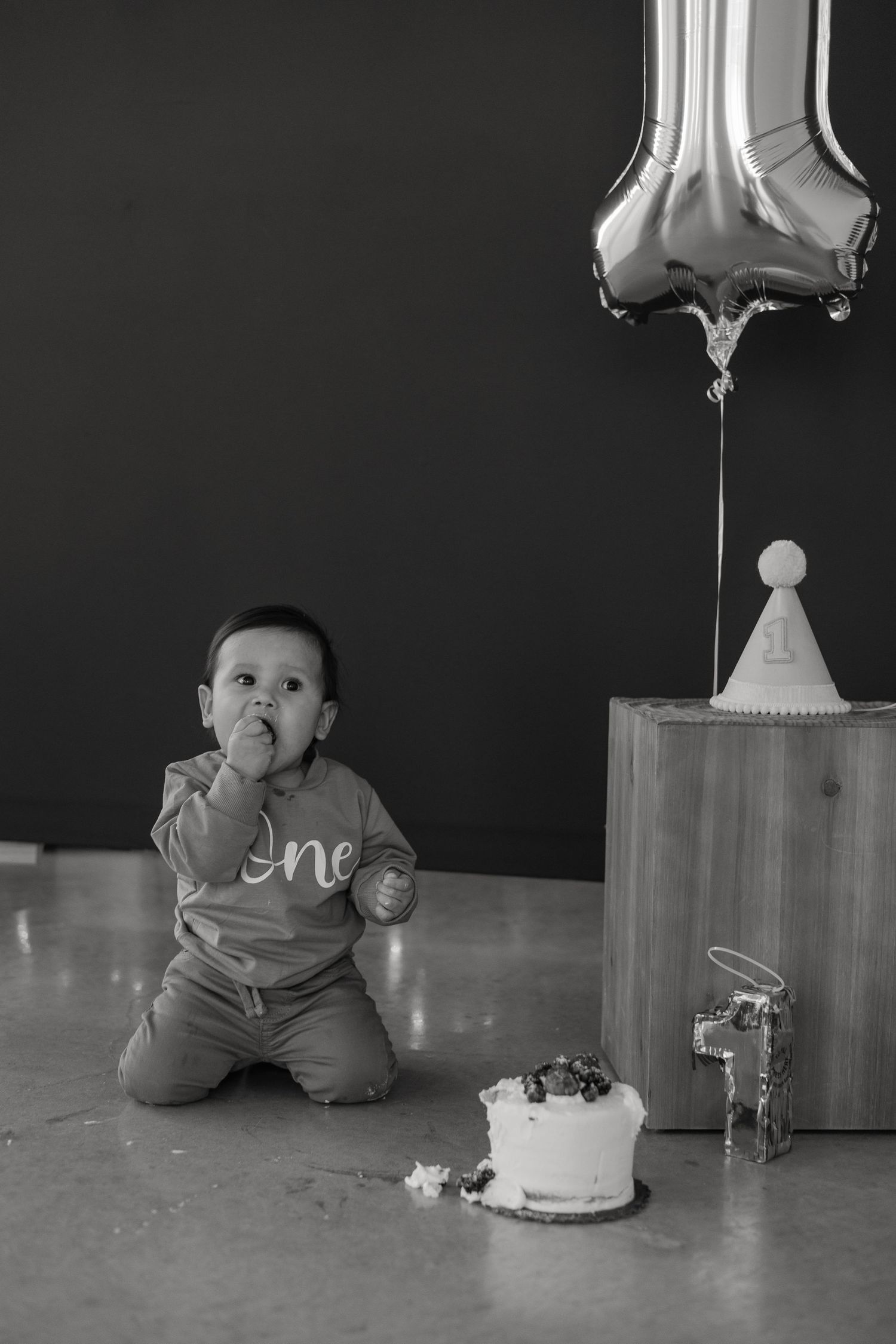 Baby in grey sweater sitting by cake and balloon in monochrome setting.