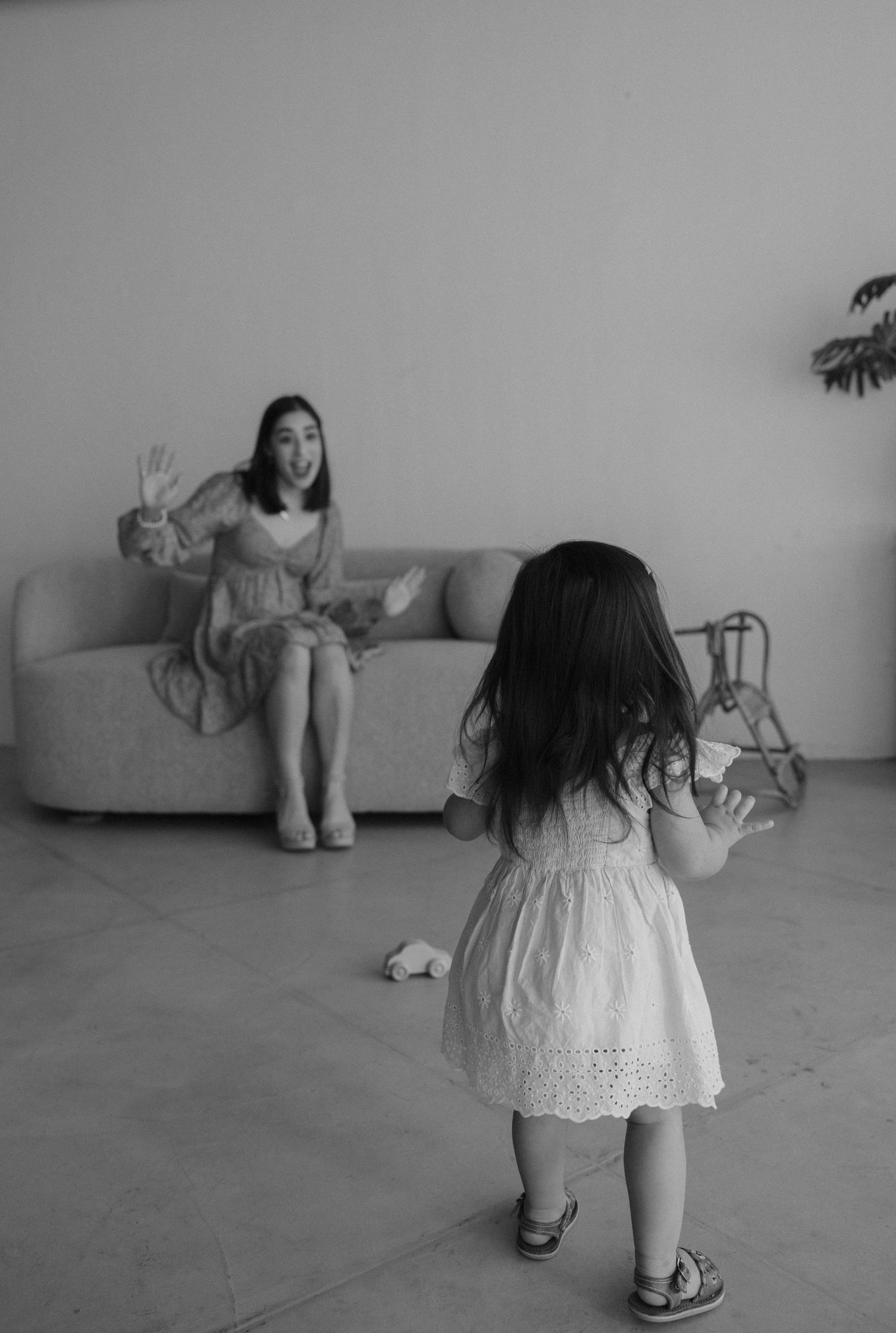 A black and white photograph showing two people in white dresses interacting in a minimalist interior space.