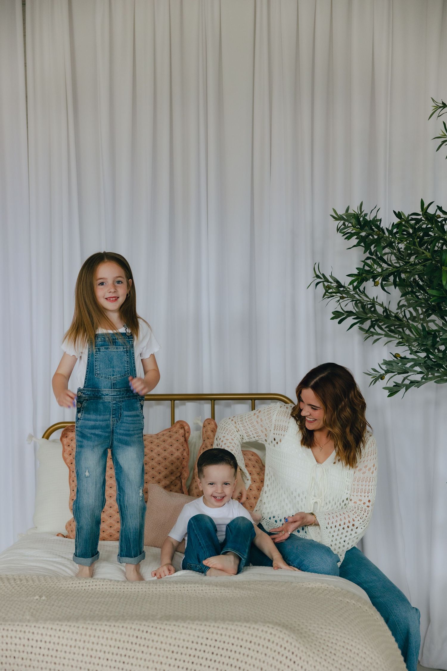 Family enjoying playful moment together on a white bed with knitted blanket next to a green potted plant.