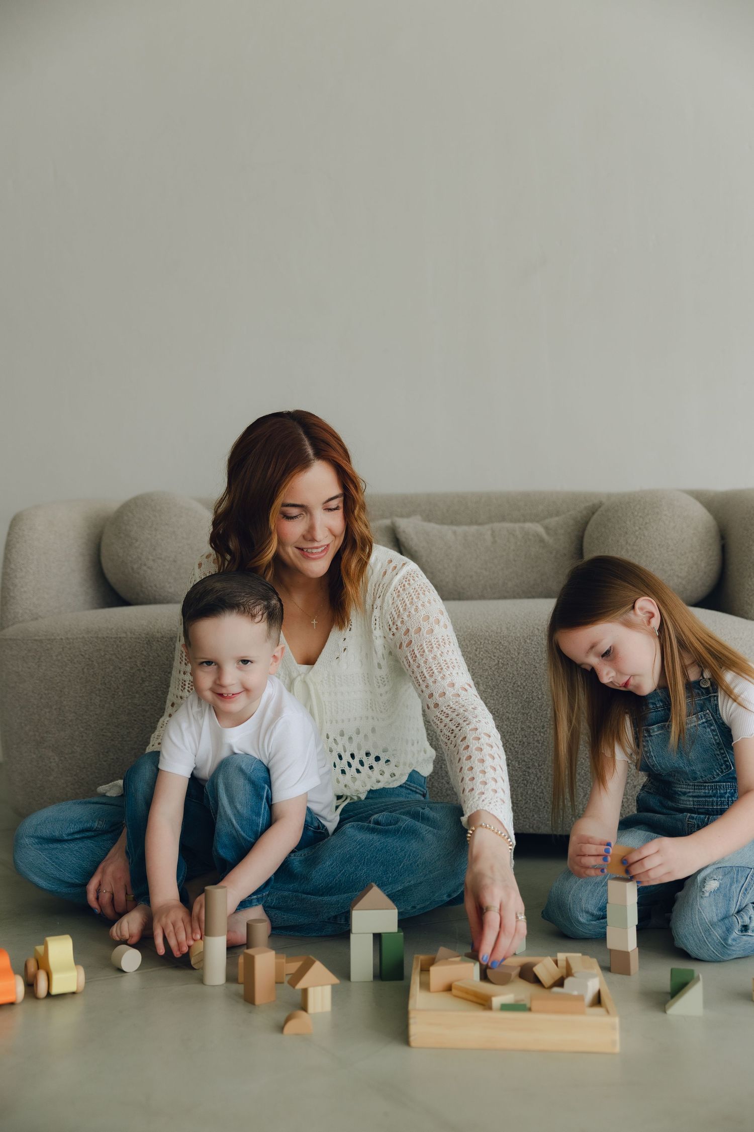 Family sitting on floor by couch playing with wooden building blocks and toys together in living room.