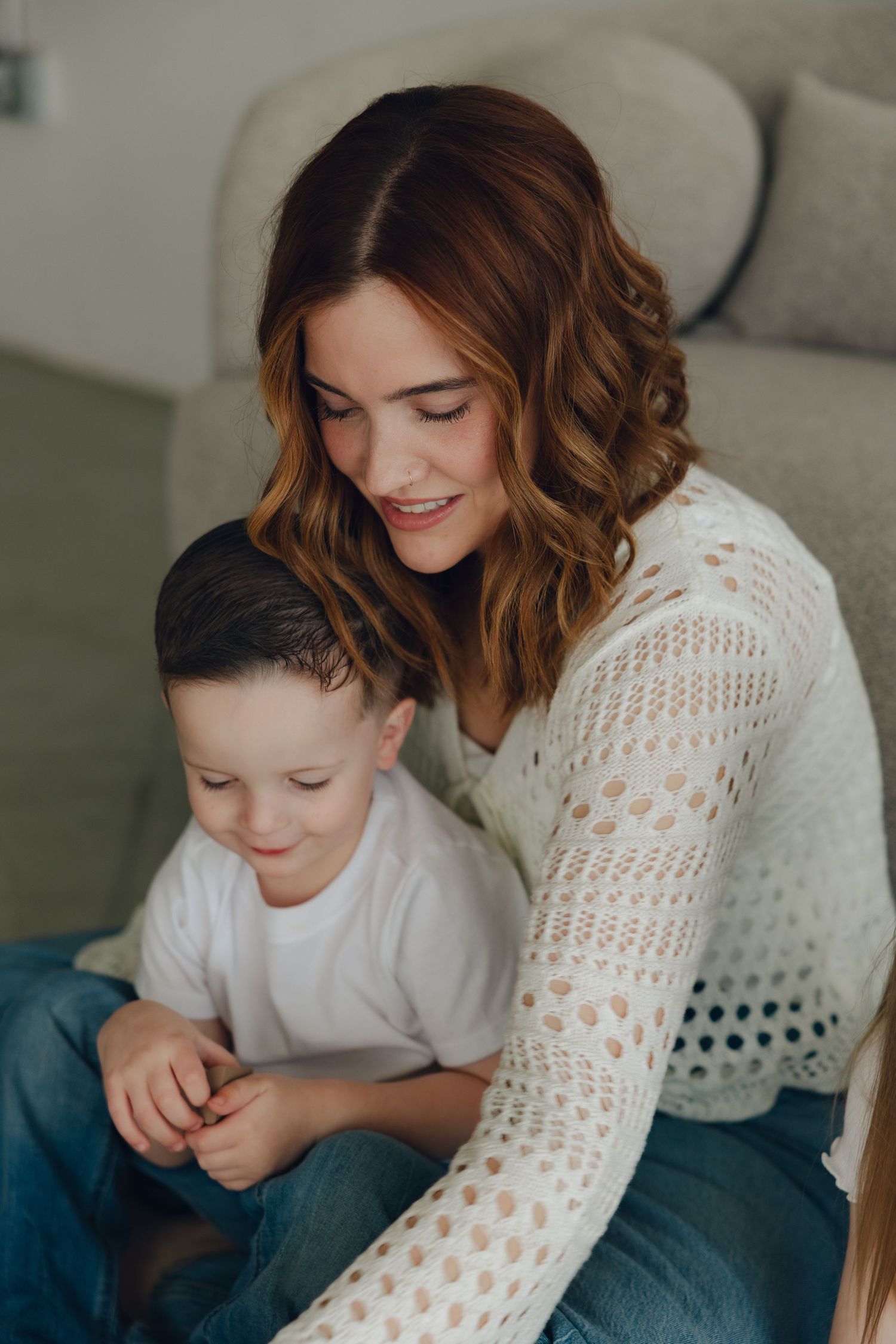 A tender moment captured as parent and child sit together on a gray couch, wearing jeans and a white crochet sweater.