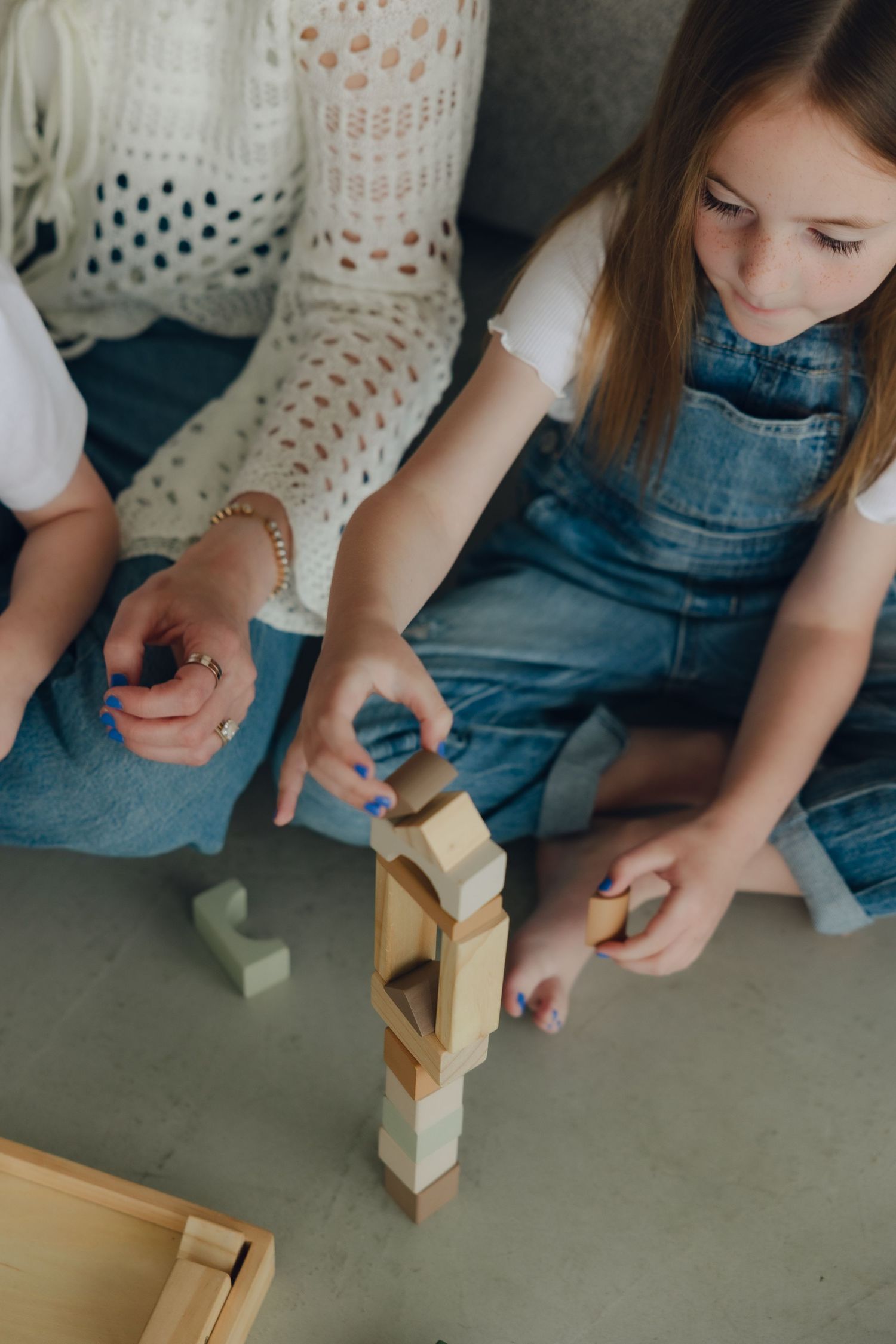 Child playing with wooden building blocks while sitting on floor in denim overalls.