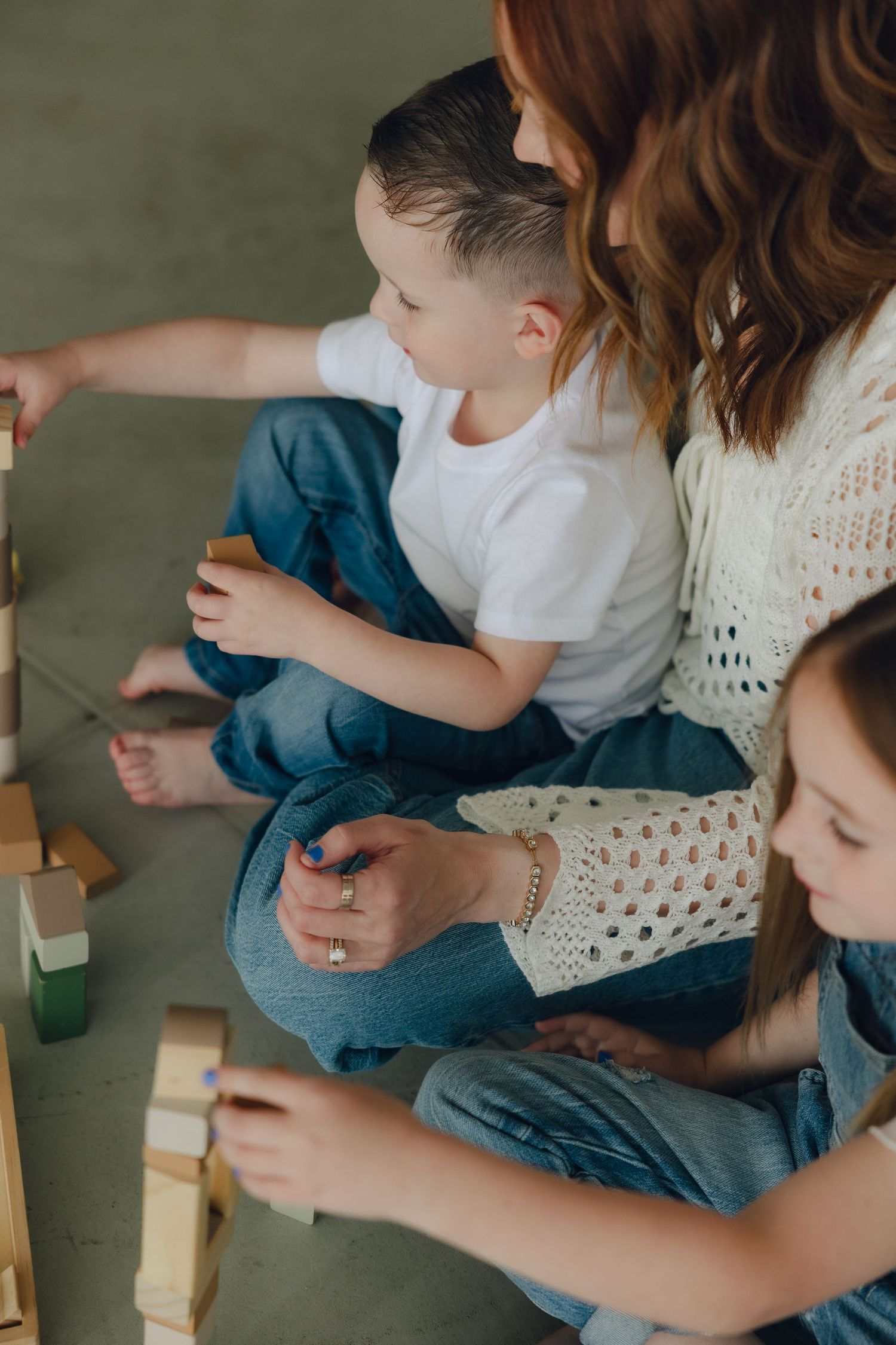 Children and adult playing with wooden building blocks while sitting on floor in white and denim casual attire.