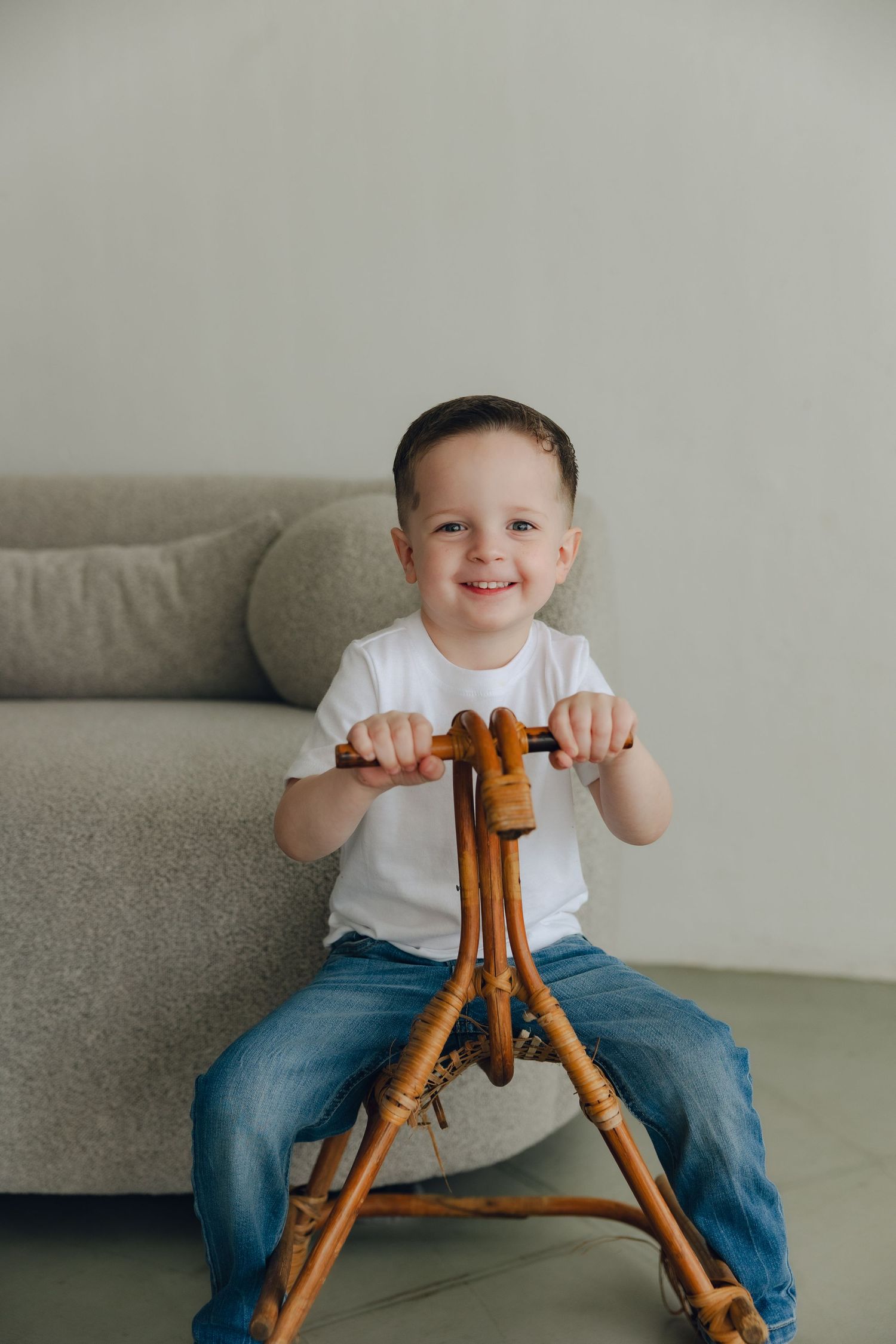 Cheerful child in white t-shirt and jeans sits on wooden rocking chair against grey couch in minimal room setting.