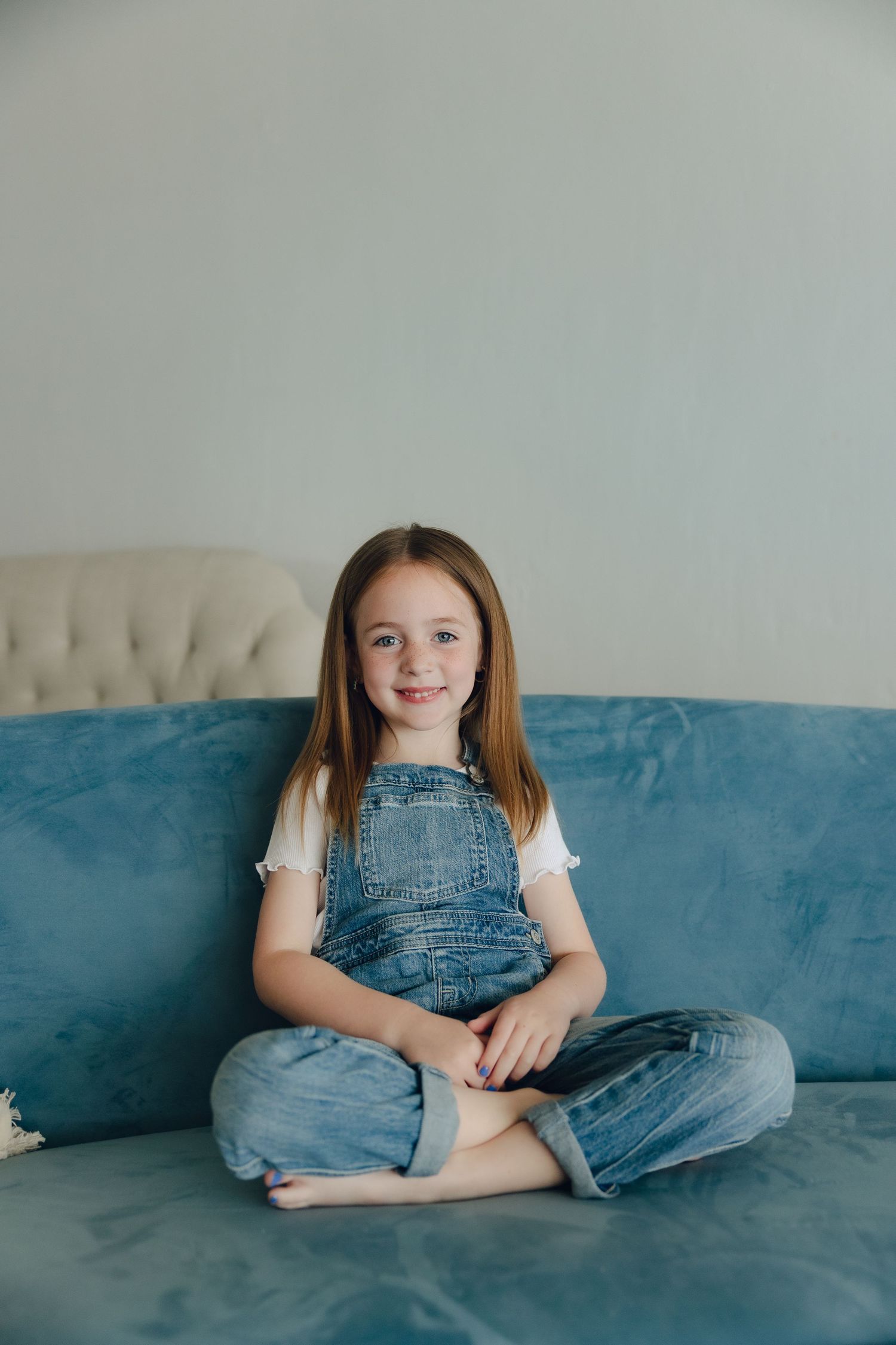 A child in denim overalls sits cross-legged on a blue velvet couch against a light gray wall.