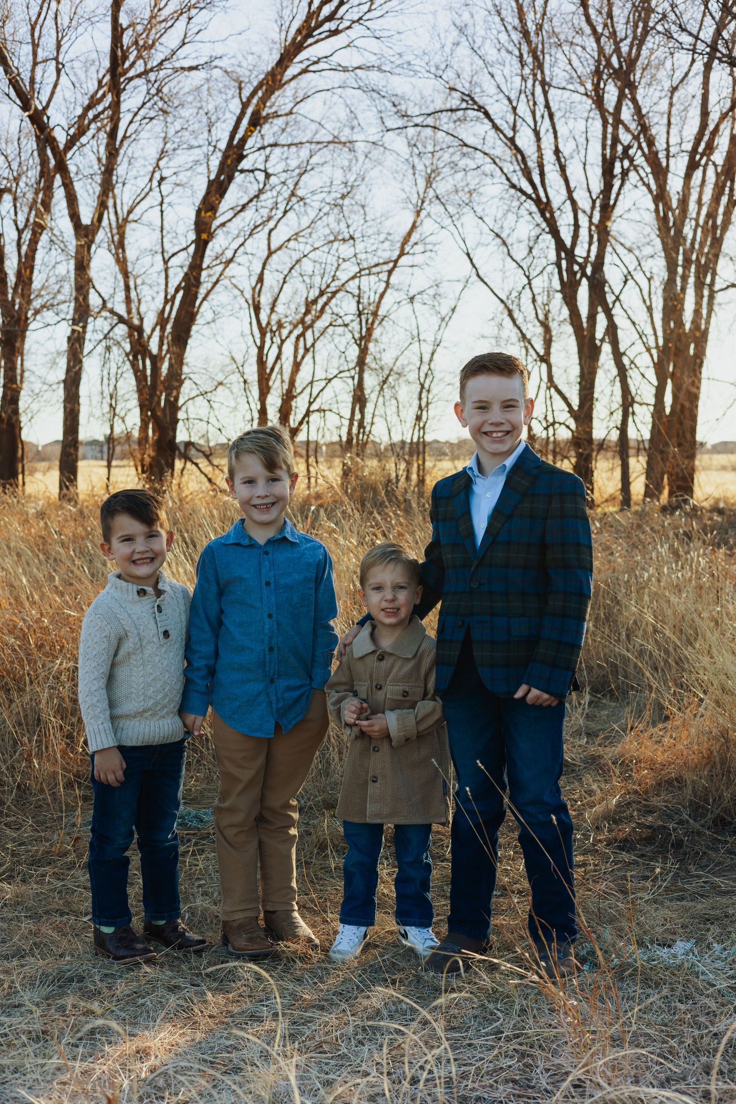 Four siblings stand together smiling in tall grass during golden hour.