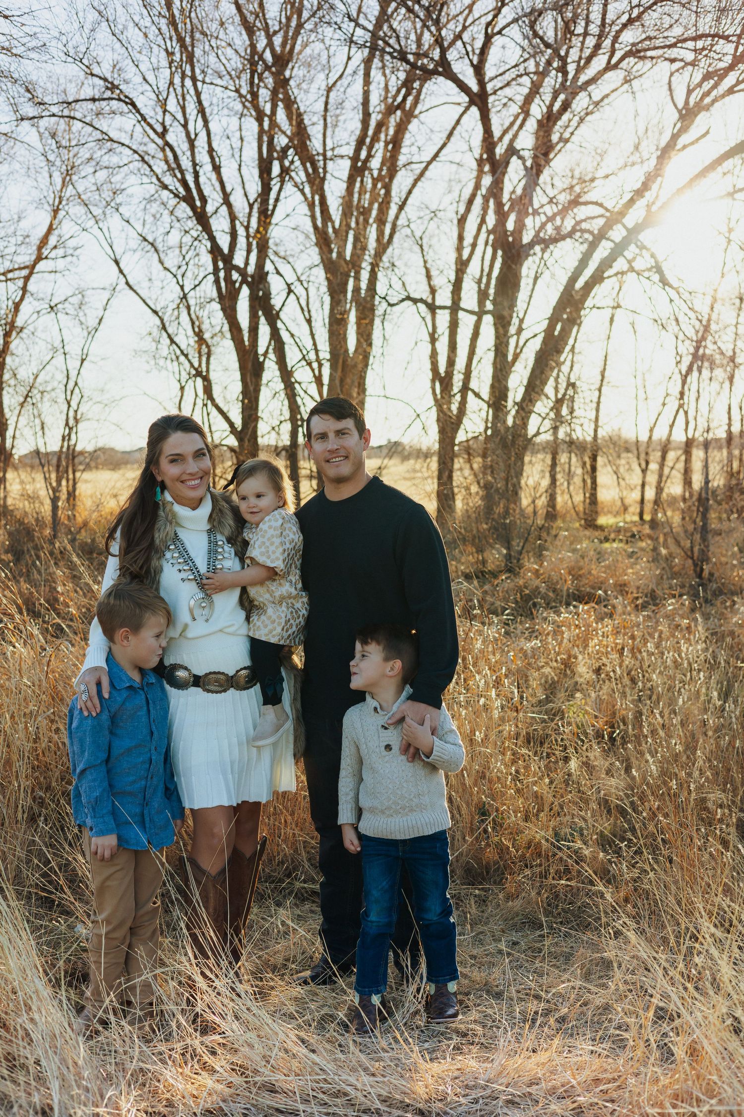 Family poses together at sunset in tall grass and bare trees.