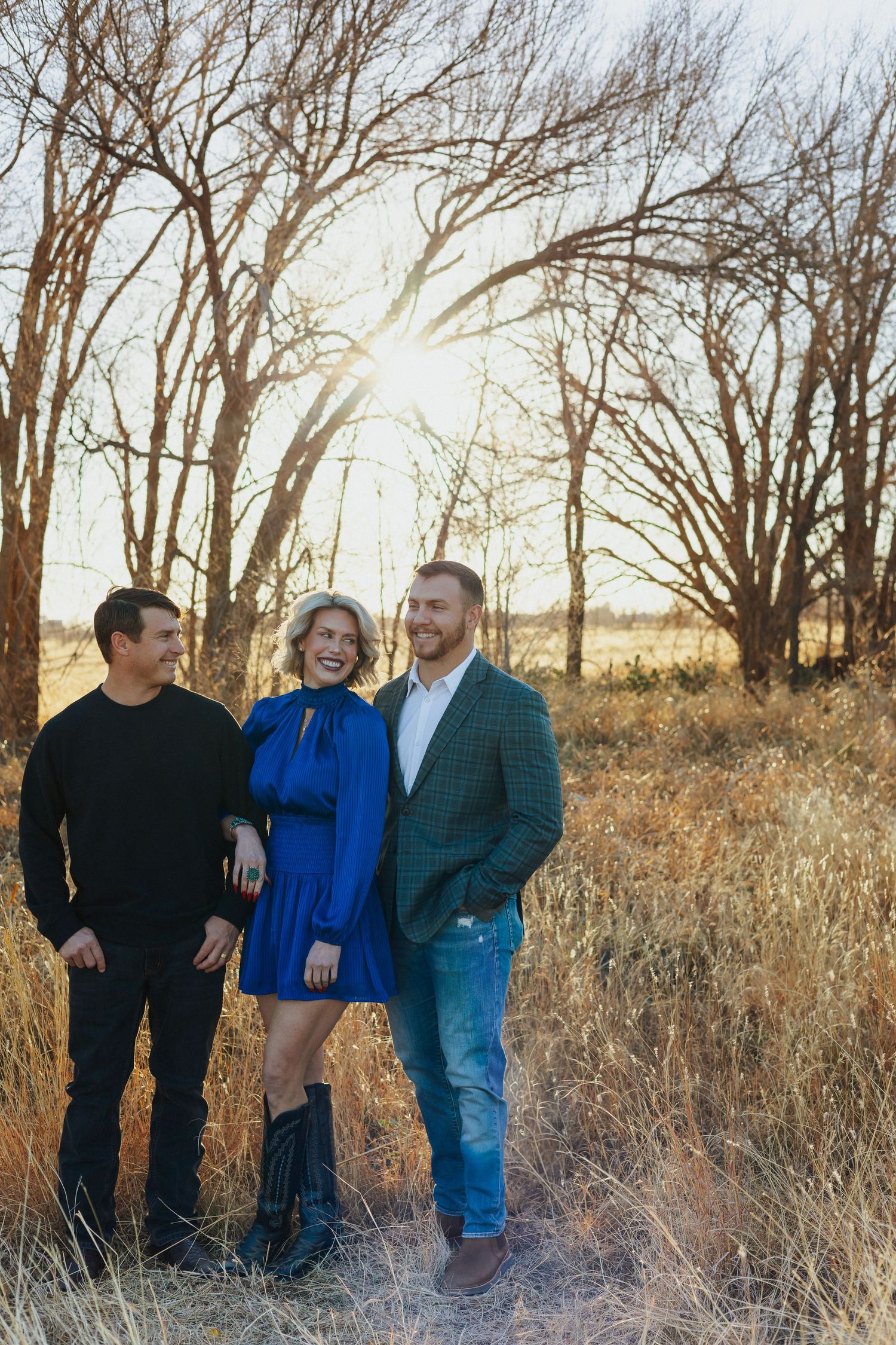 Three friends smile together in a field at sunset with bare trees.