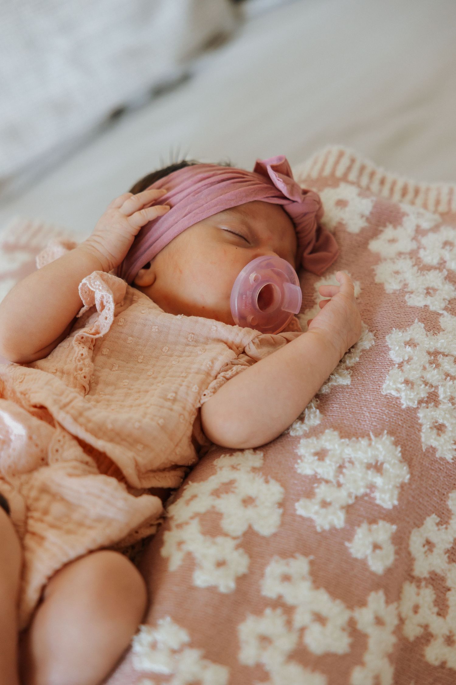 Sleeping newborn wearing pink headband and peachy outfit on floral blanket.