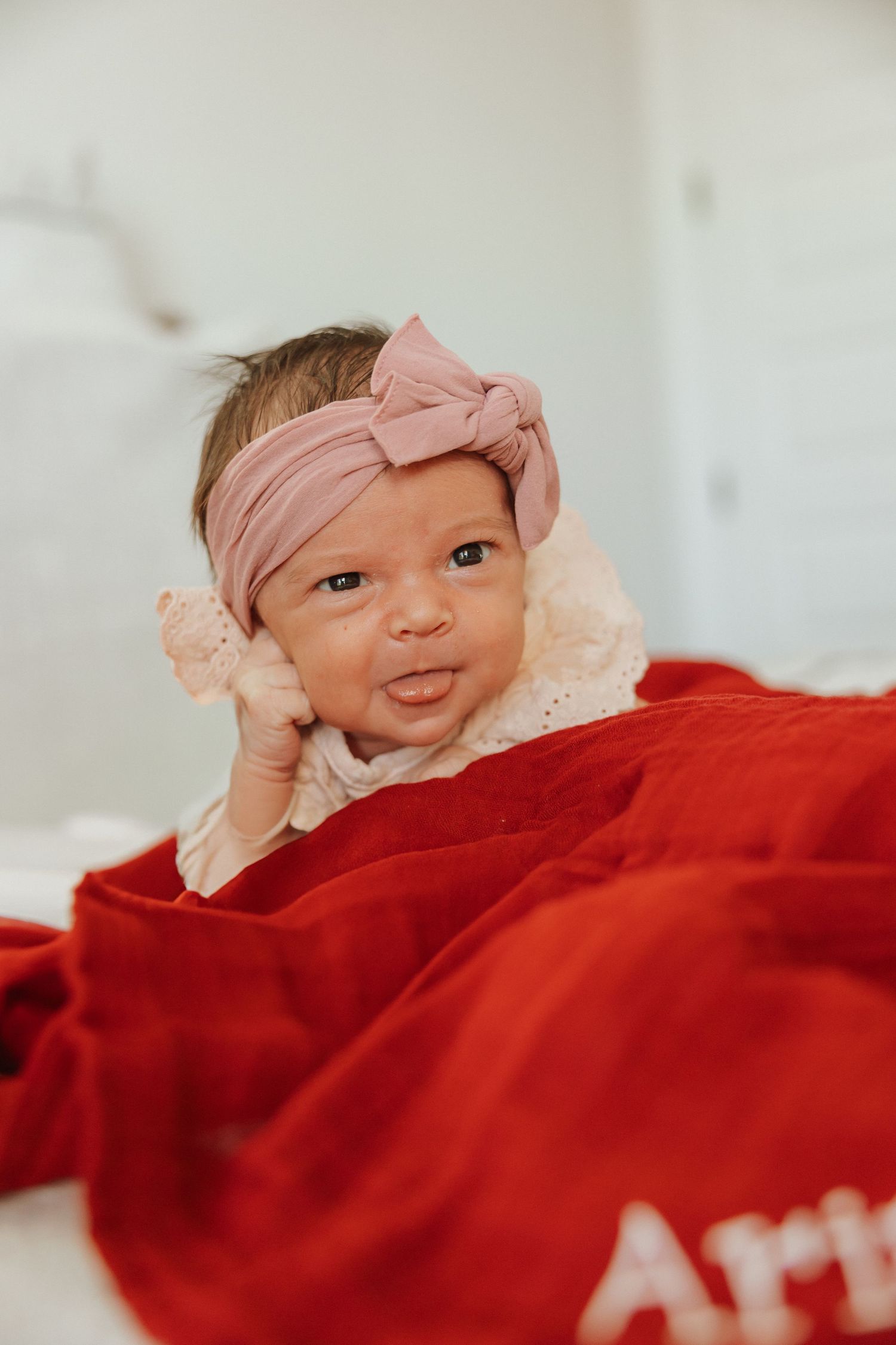 Baby wearing pink bow headband smiling while laying on red blanket.