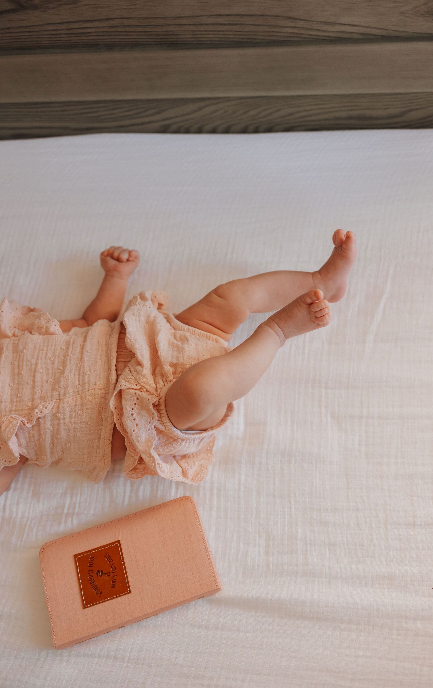 Baby legs in peachy romper on white bedding with phone beside.