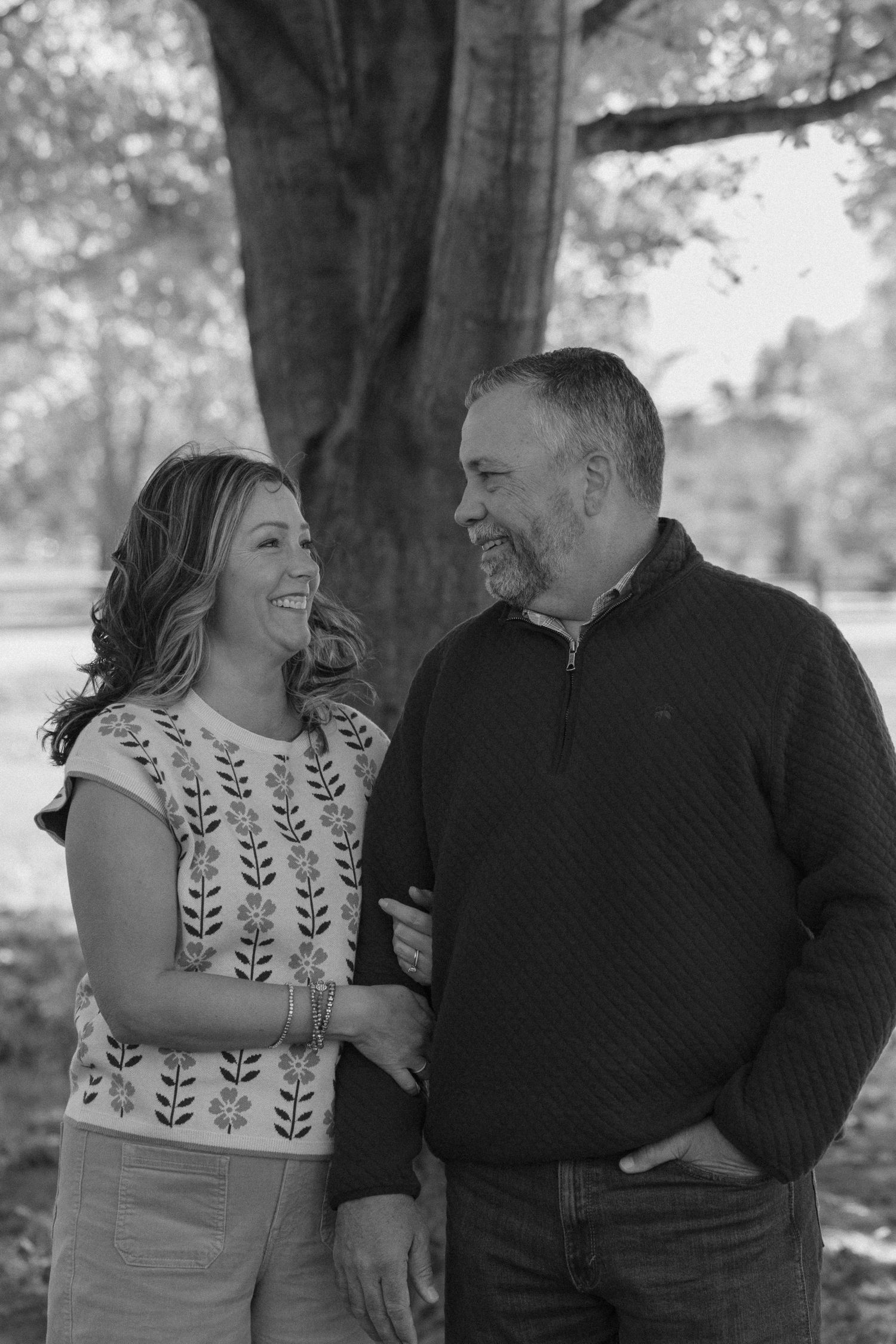 A couple shares joyful laughter together during an outdoor black and white photo session in a park setting.
