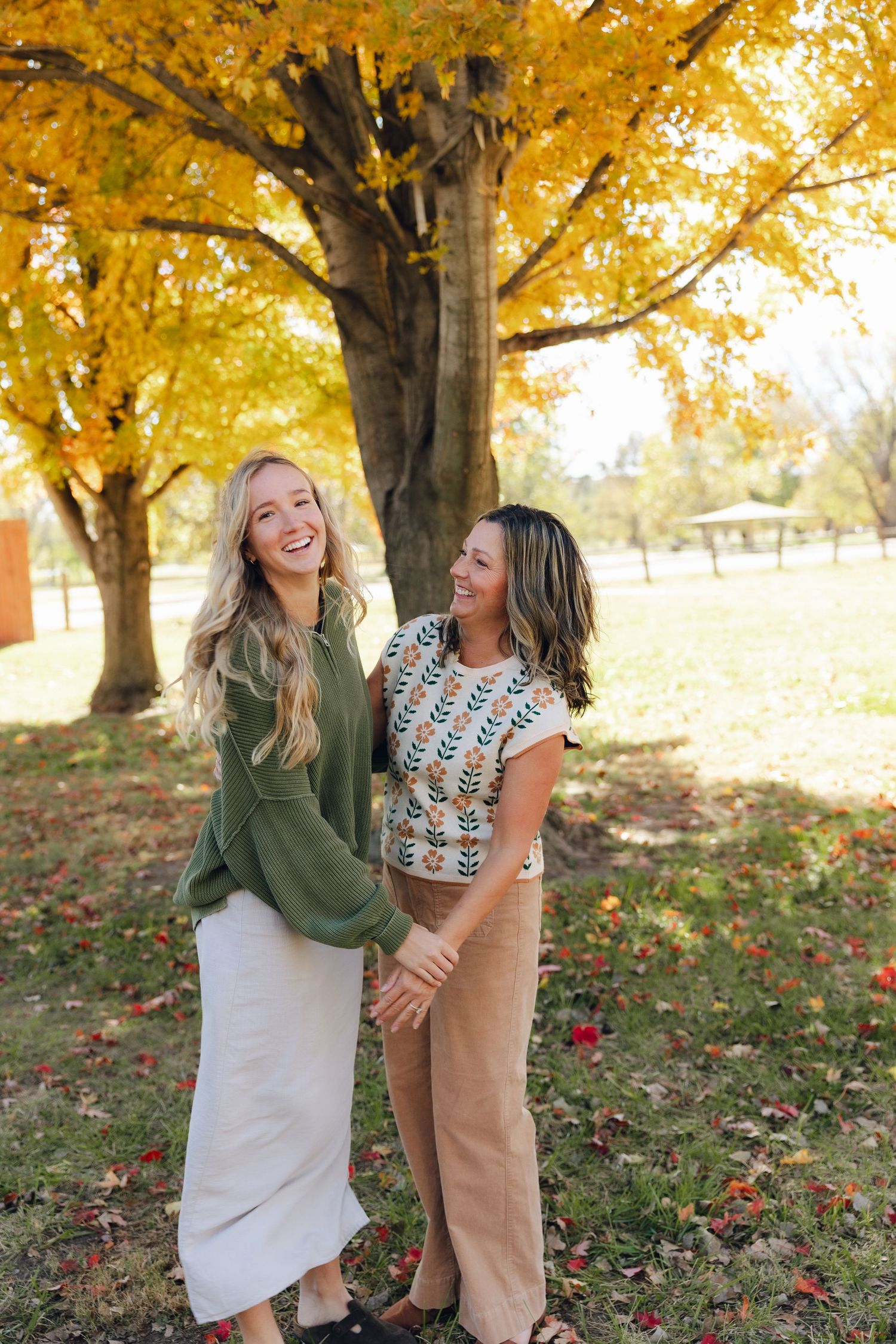 Two friends laugh together while enjoying a beautiful autumn day with golden trees and fallen leaves in the background.