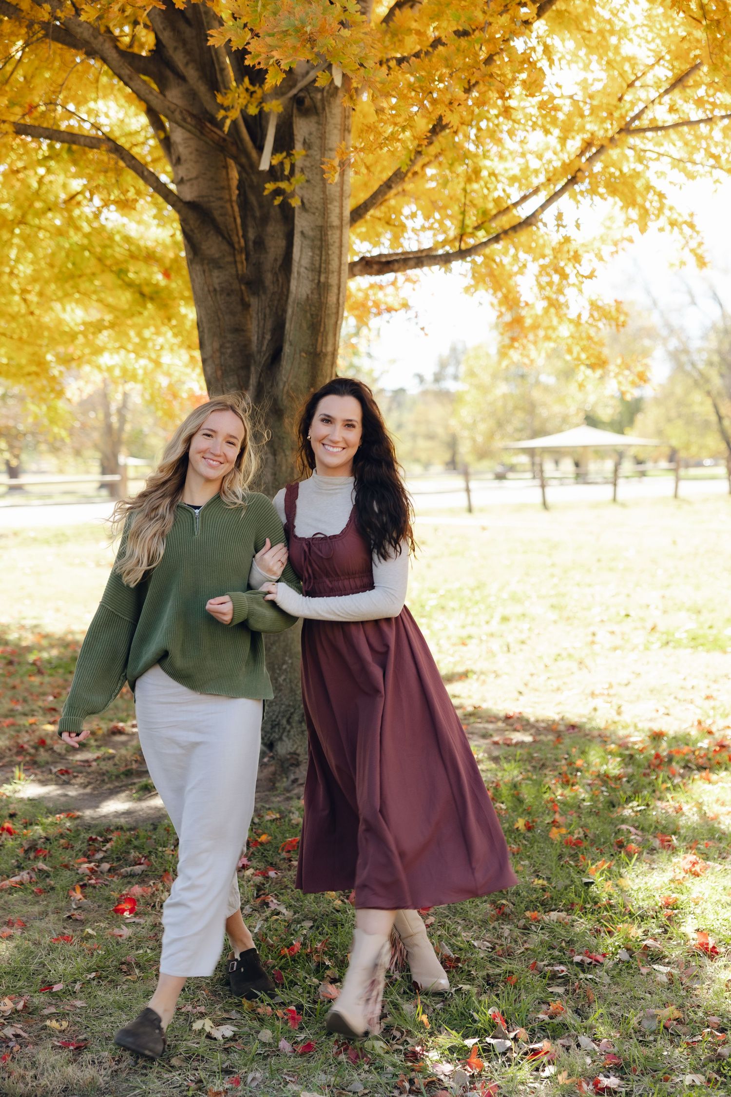 Two people pose together in autumn setting with golden yellow leaves and trees in background.