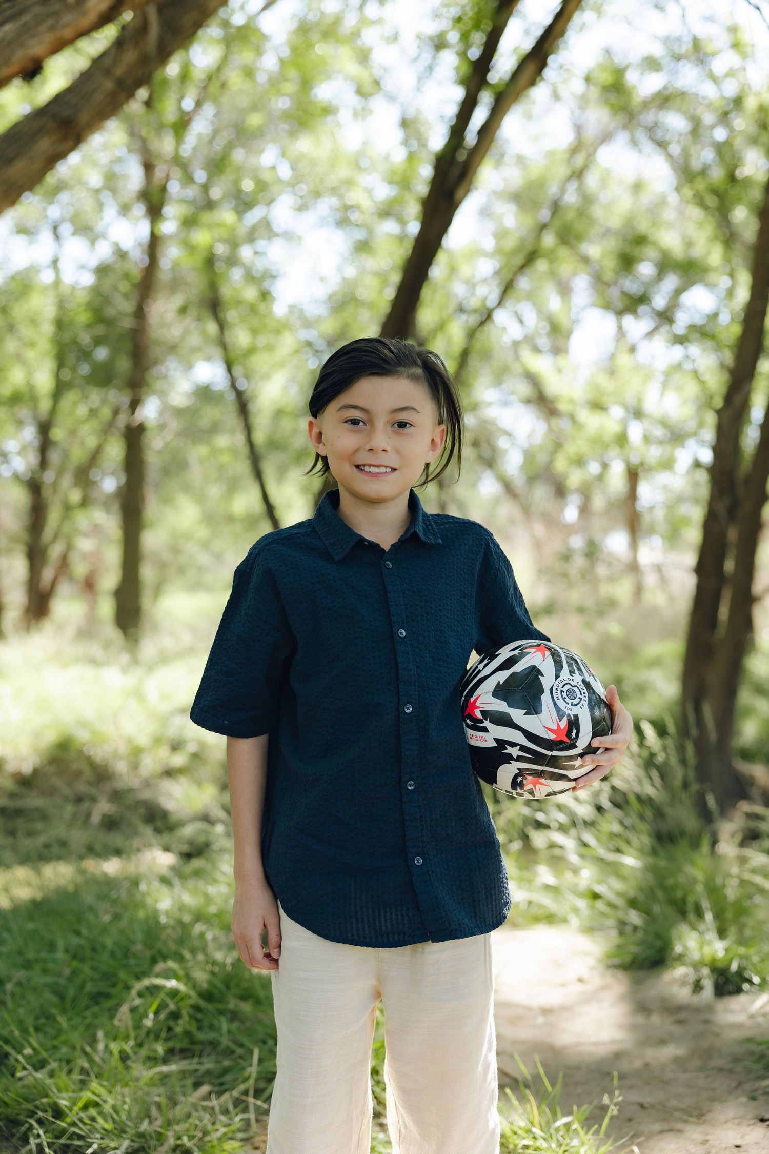 Youth wearing navy shirt and white pants holds soccer ball while standing on forest path in natural outdoor setting.