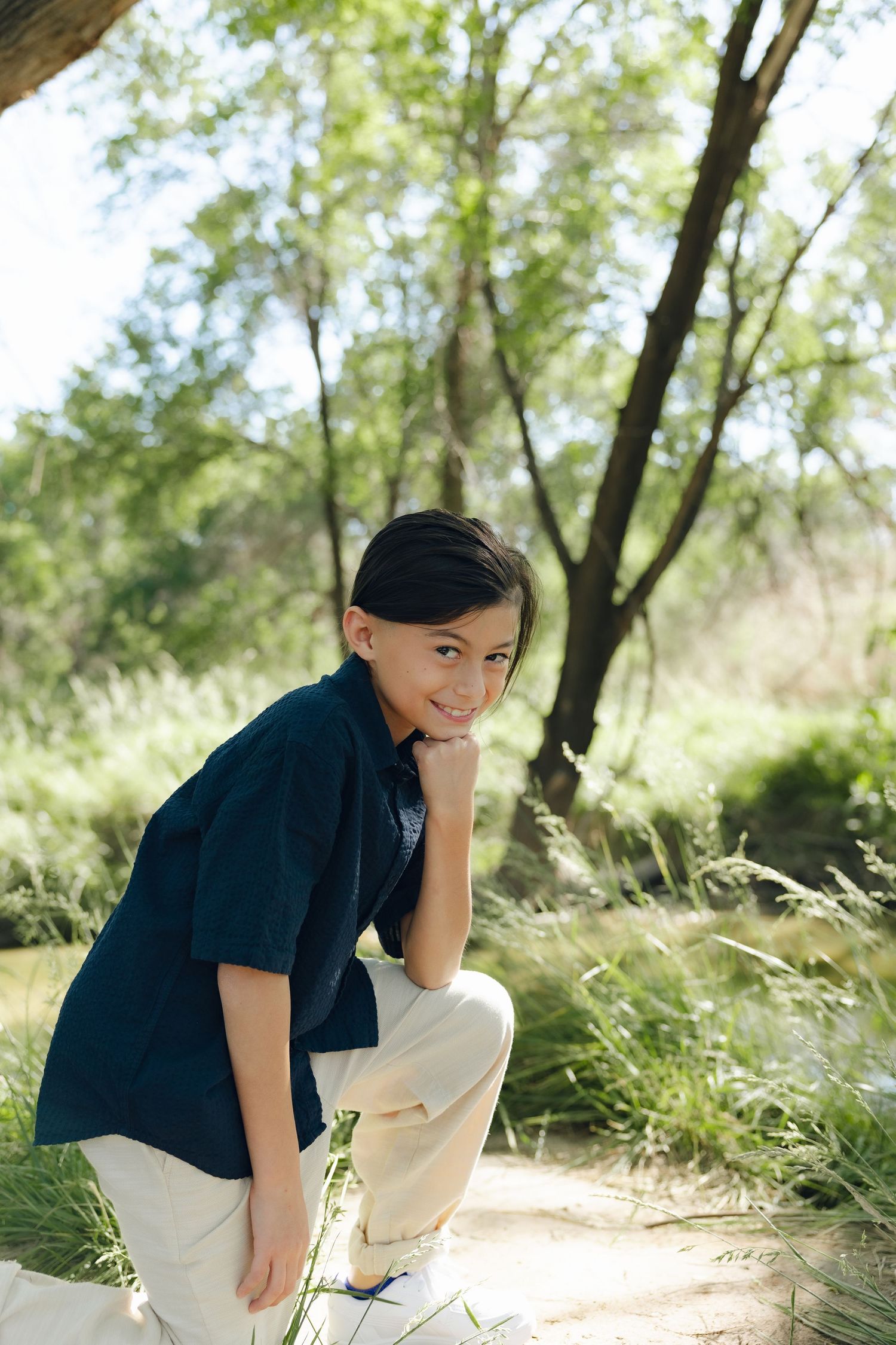 Child in navy shirt and khaki pants poses thoughtfully in a sunny outdoor setting surrounded by trees and grass.