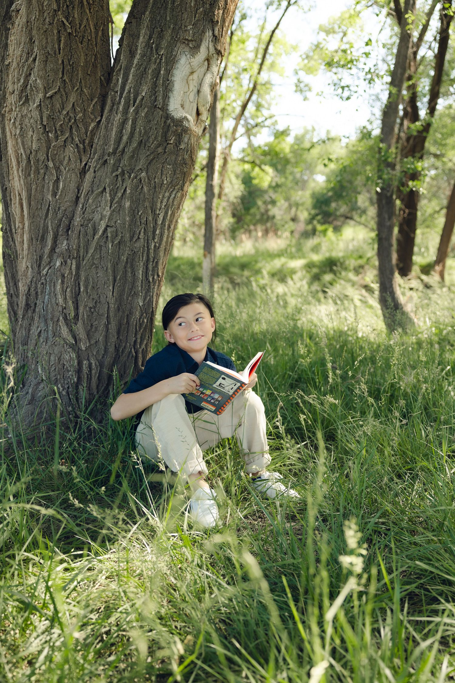 Child reading a book while sitting against a large tree trunk in a peaceful natural setting with tall grass.