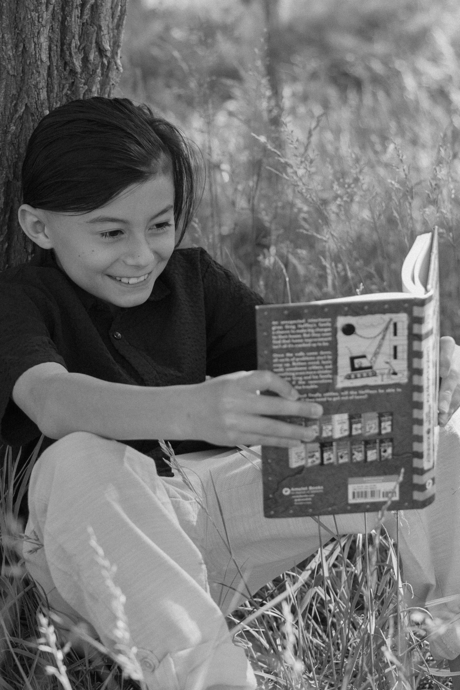 A black and white portrait of a young person reading a book while sitting in tall grass against a tree trunk.