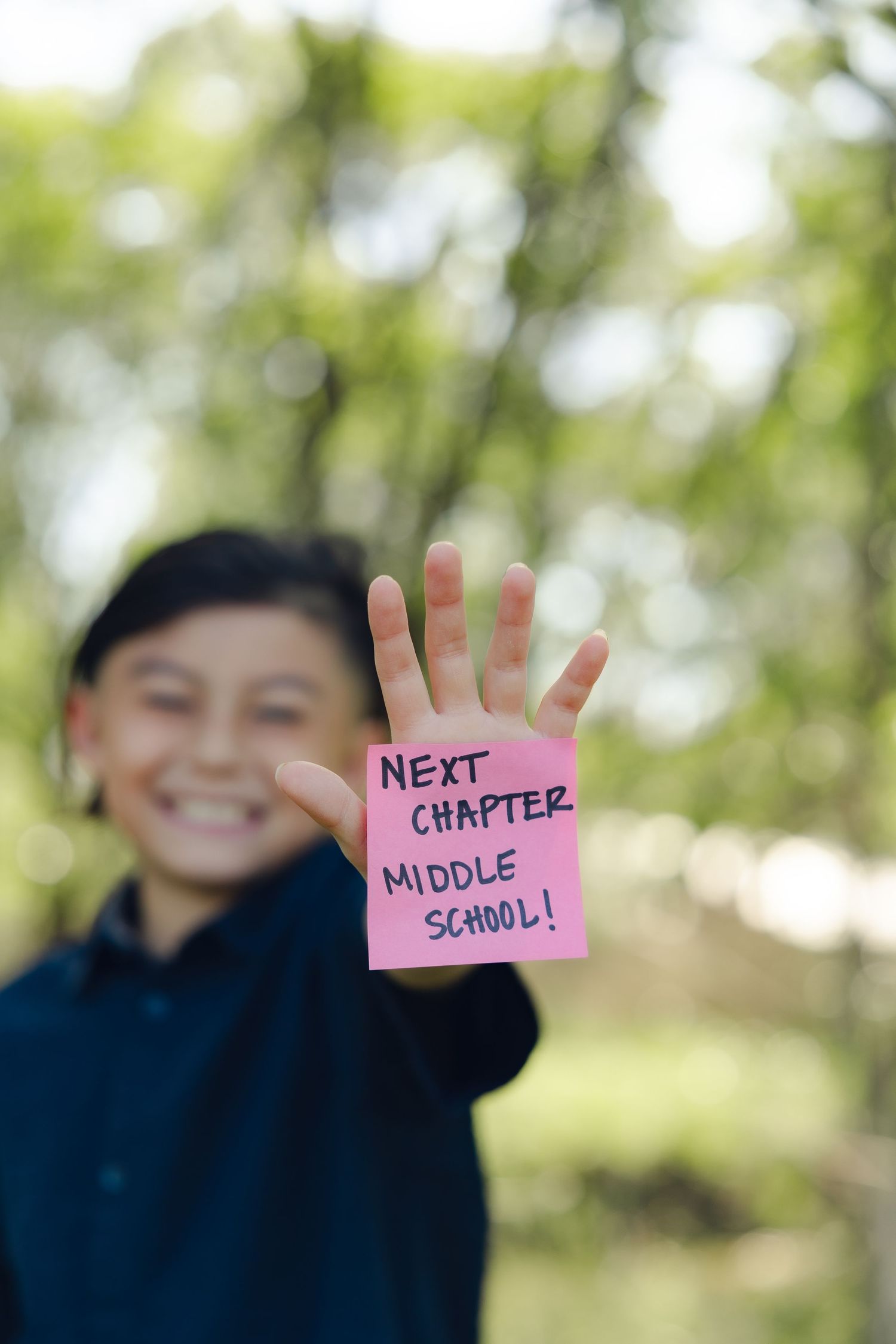 Hand holding up a pink sticky note with "Next Chapter Middle School" written on it against a blurred outdoor background.