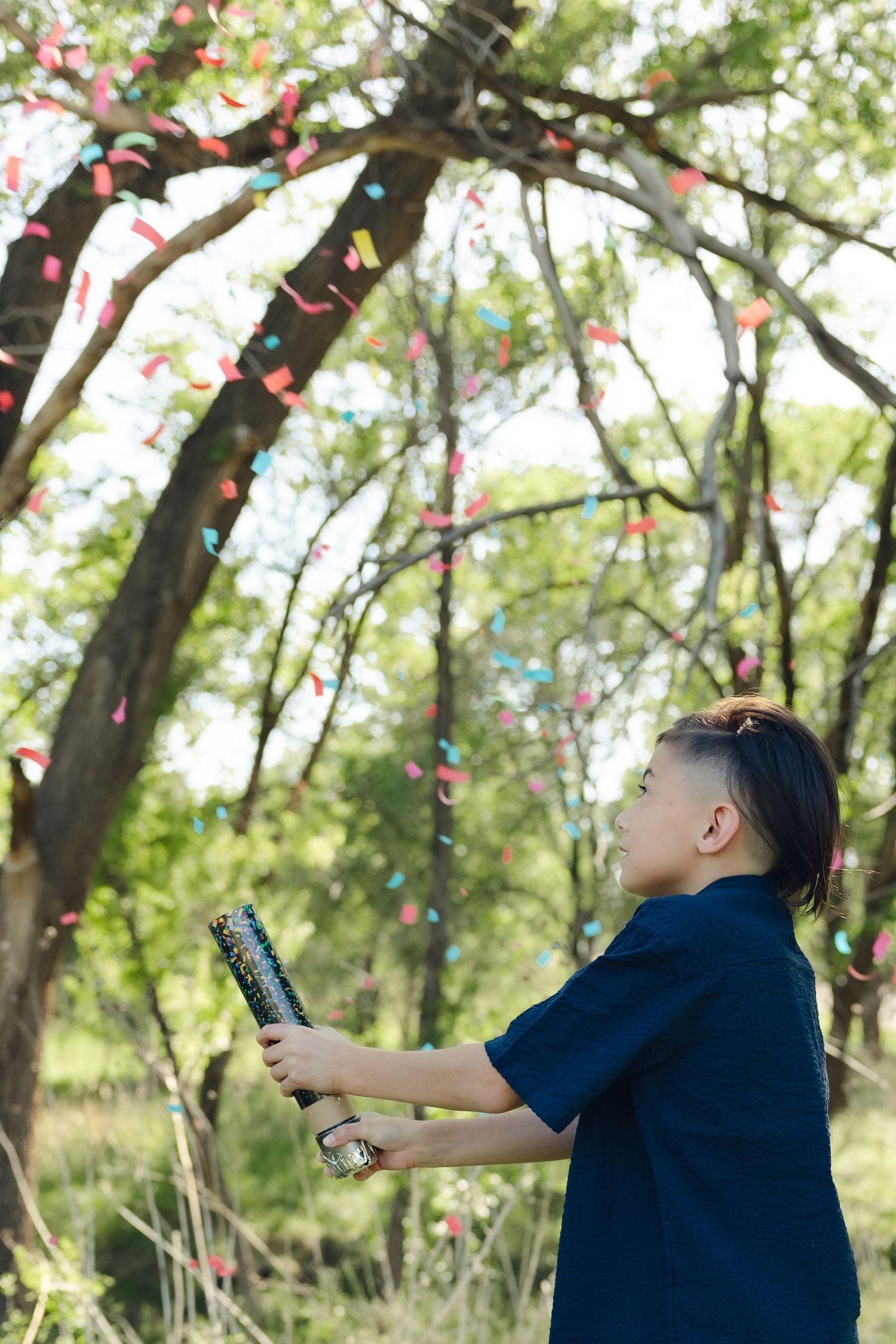 Person in navy blue polo shirt opening a confetti popper outdoors with colorful confetti falling through tree branches.