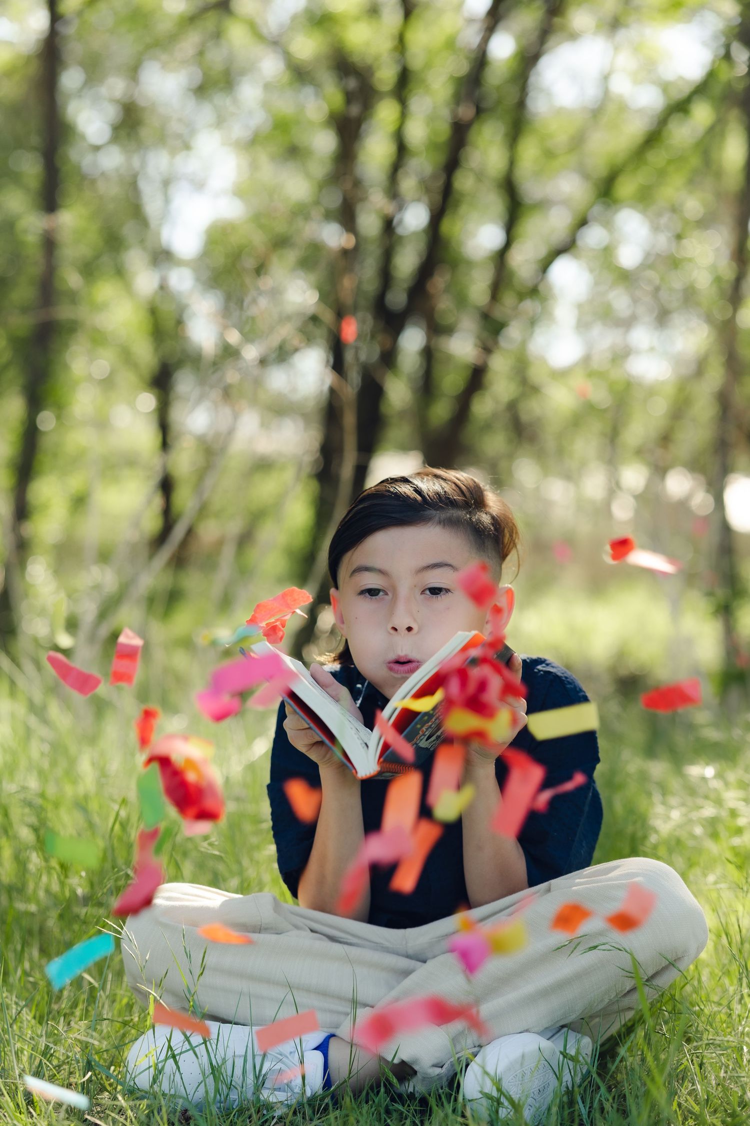 Child sitting cross-legged on grass blowing red flower petals in a sunlit forest setting.