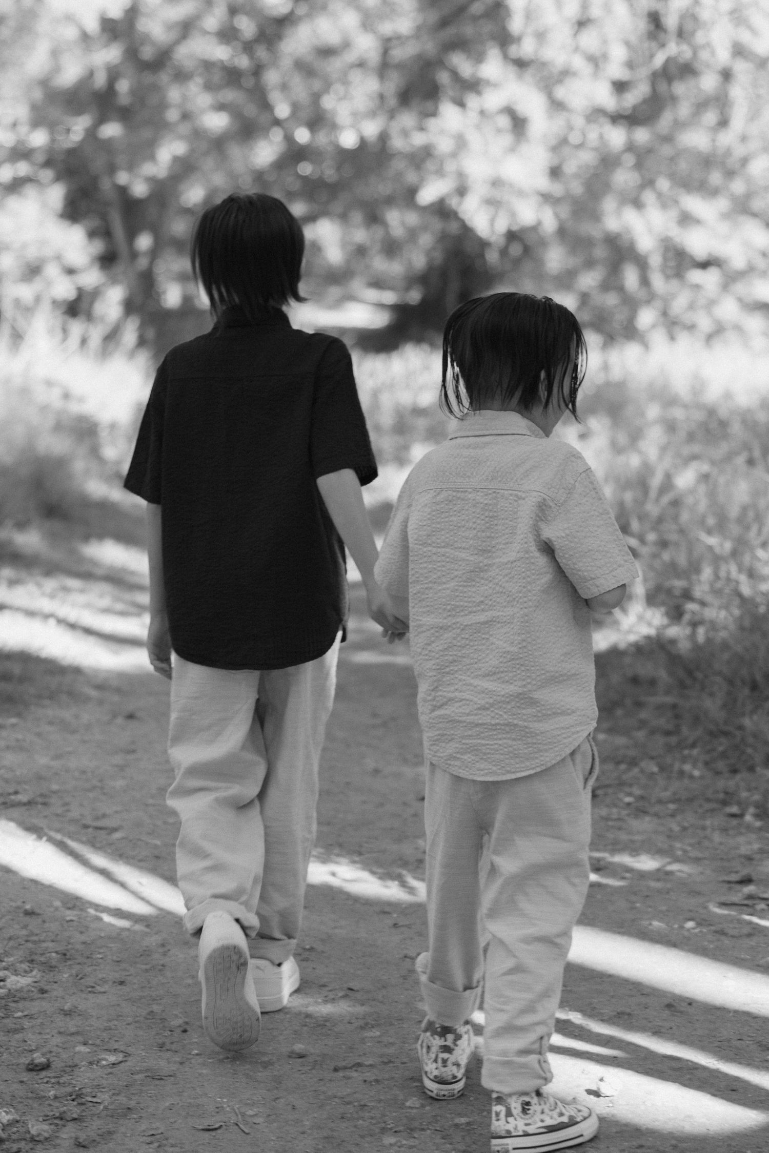 Back view of two children walking together on a dirt path through a sunlit wooded area.