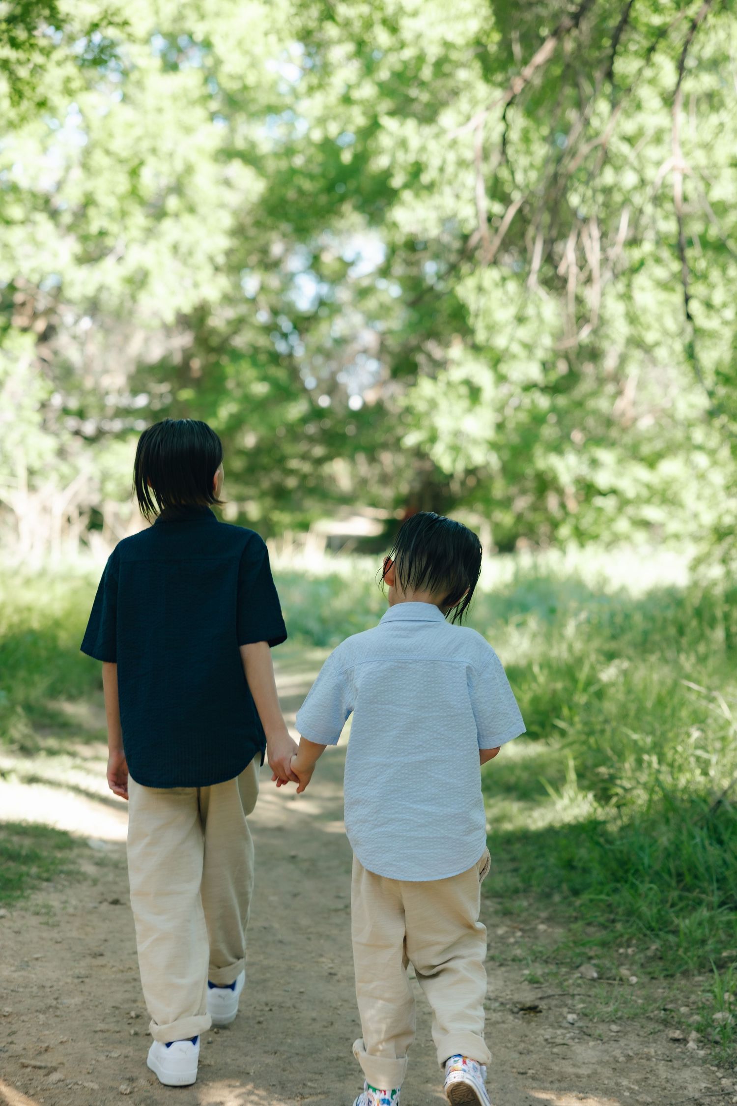 Two young siblings walking hand in hand down a sunlit nature trail in summer attire.