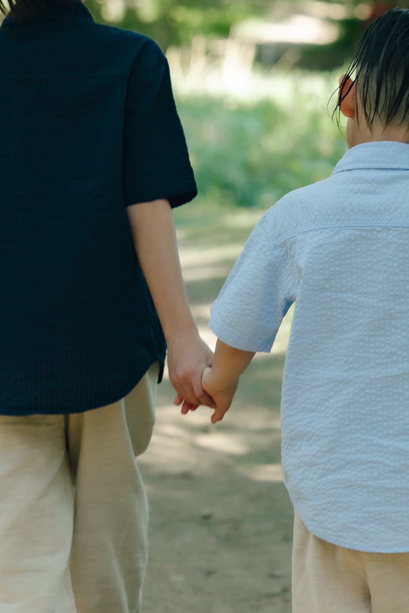 Two people in casual white and navy shirts holding hands while walking together on a dirt path on a sunny day.