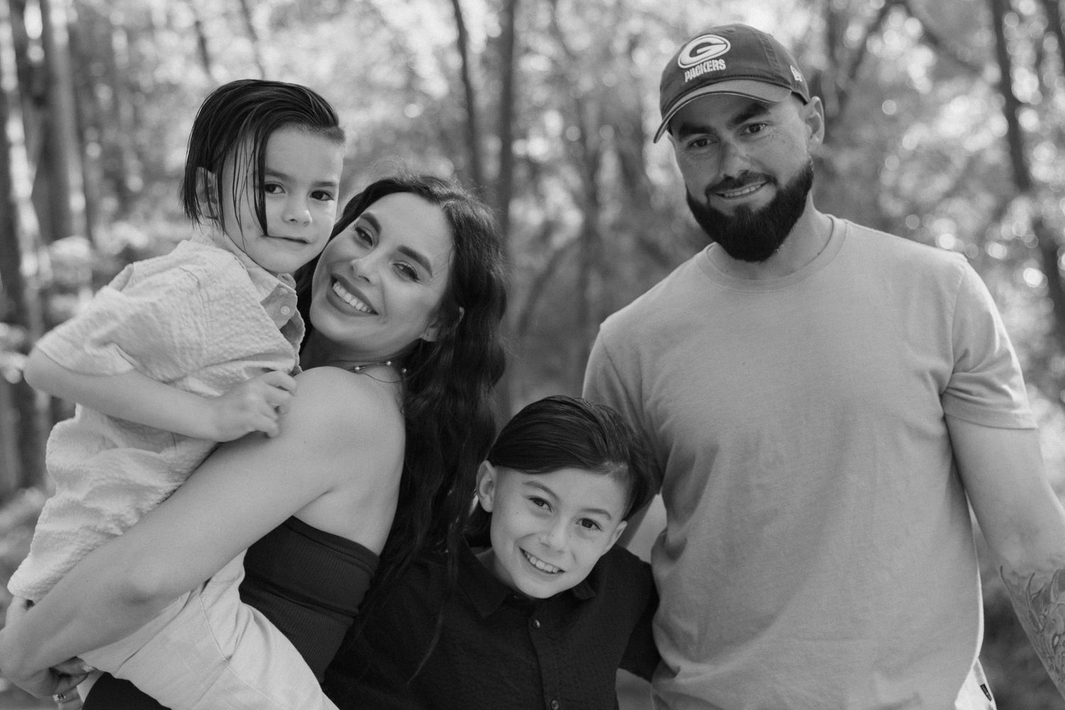 A black and white family portrait taken outdoors among trees shows a loving family of four together.