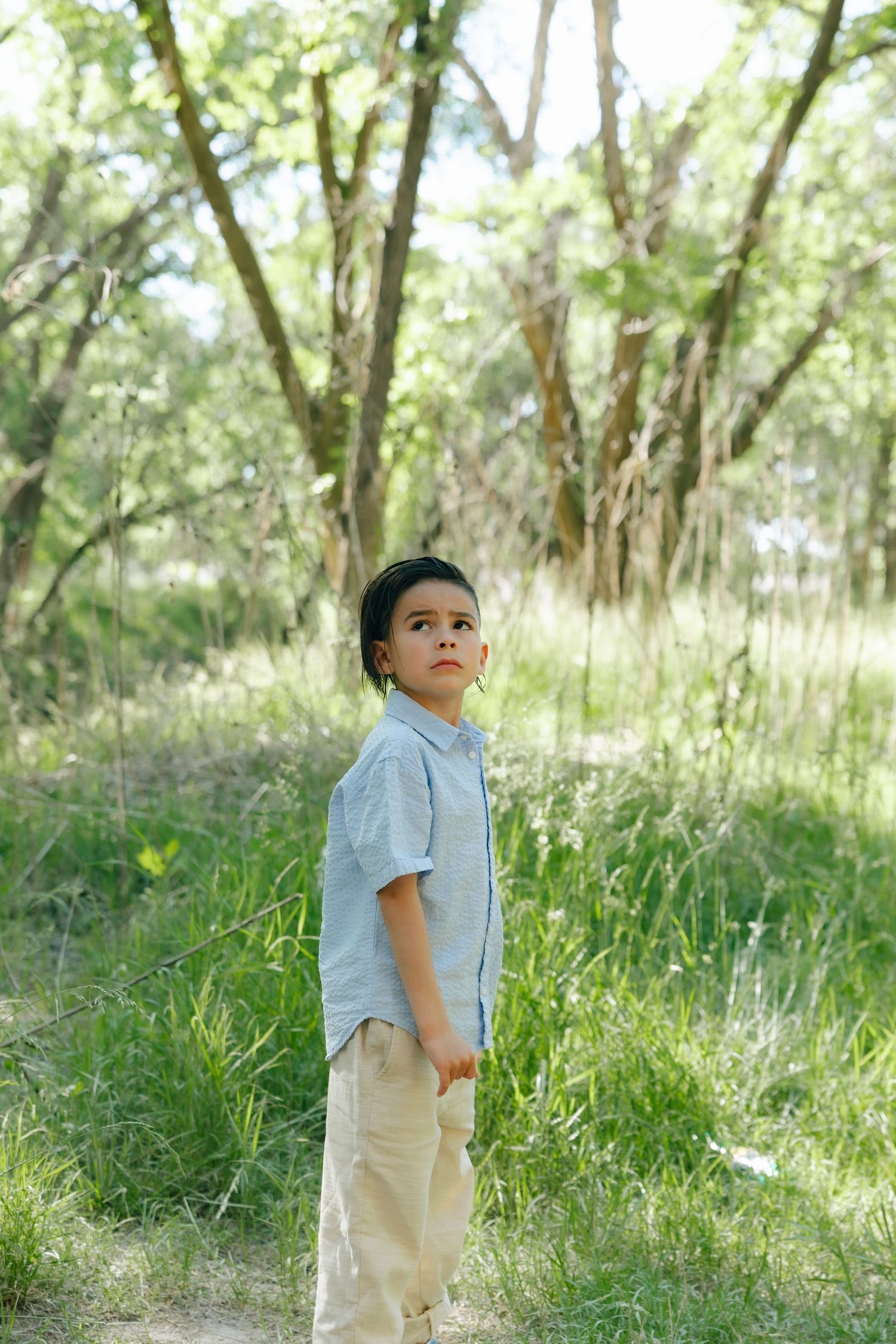 Young person in light clothing standing thoughtfully on a nature path surrounded by green trees and grass.