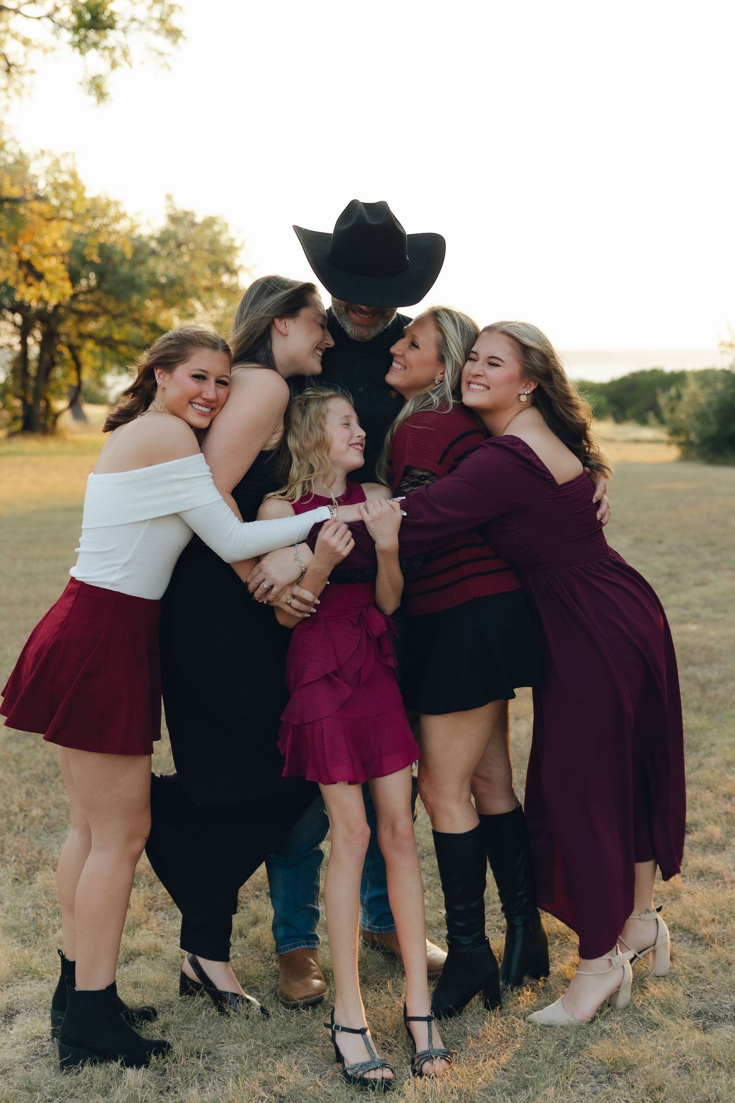A group in burgundy dresses and black attire huddle together for a candid photo shoot in a sunlit outdoor field.