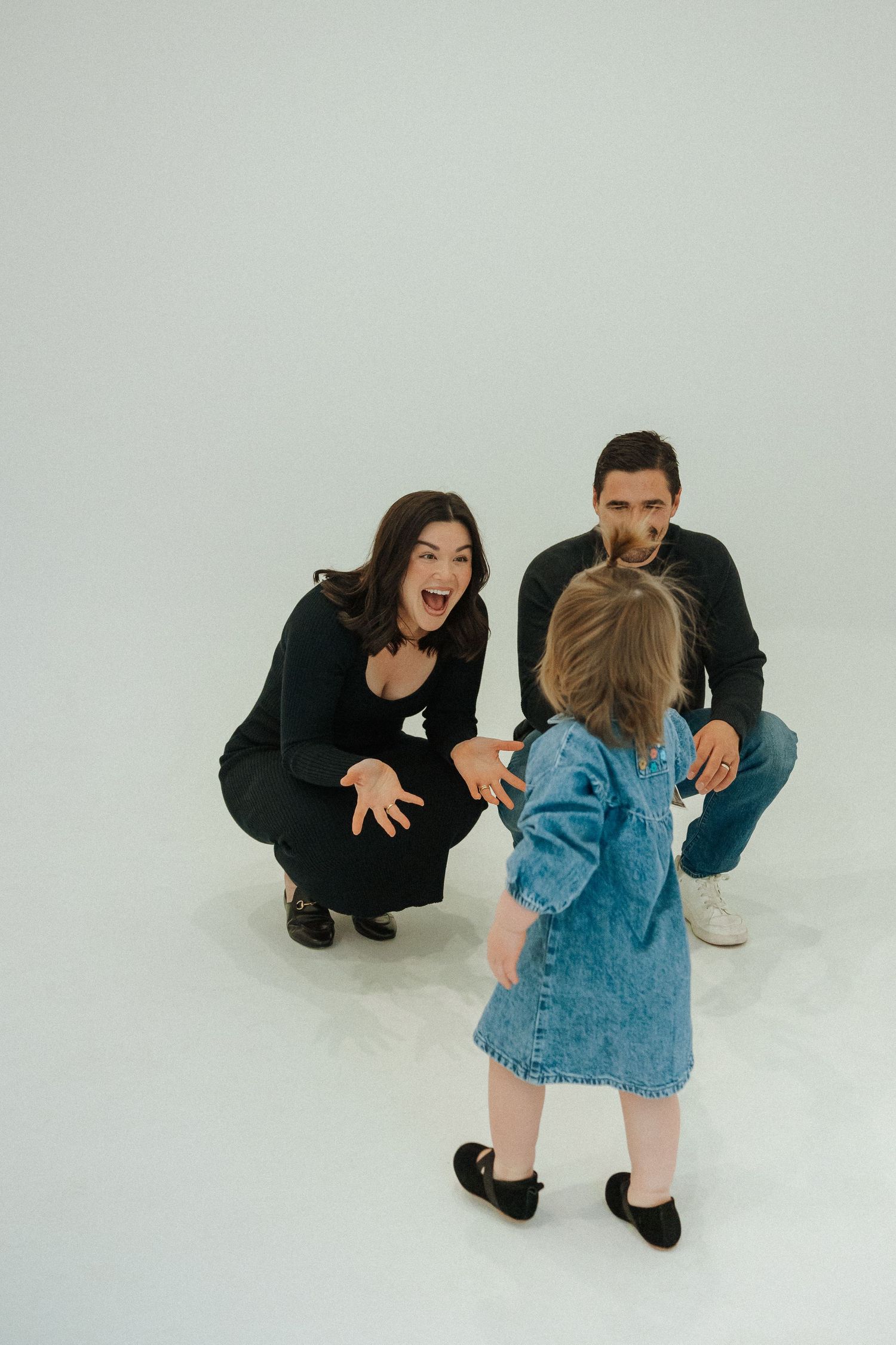 Family interacting playfully with toddler in denim dress against white background.