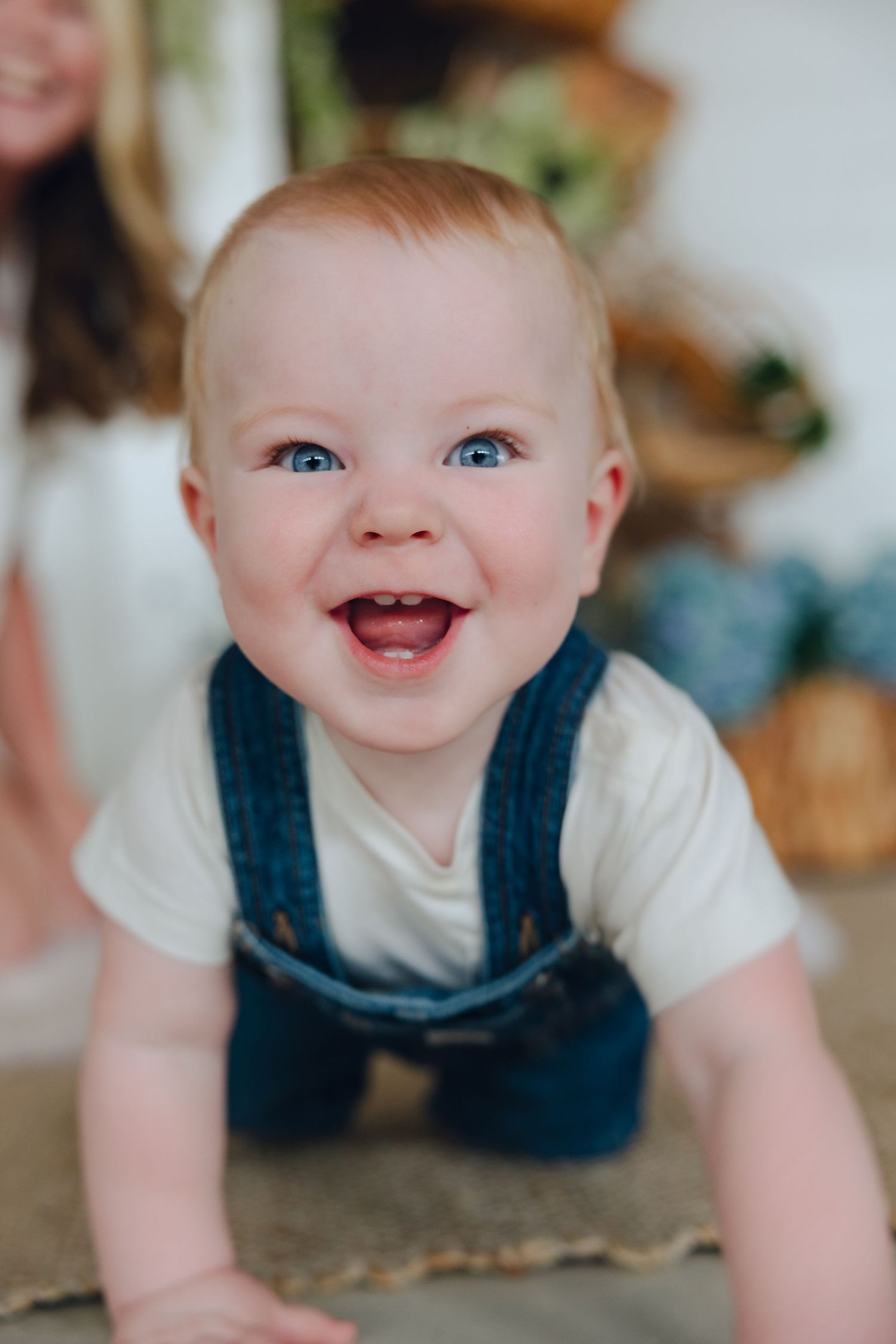 Baby wearing denim overalls and white shirt smiles with bright eyes and delighted expression.