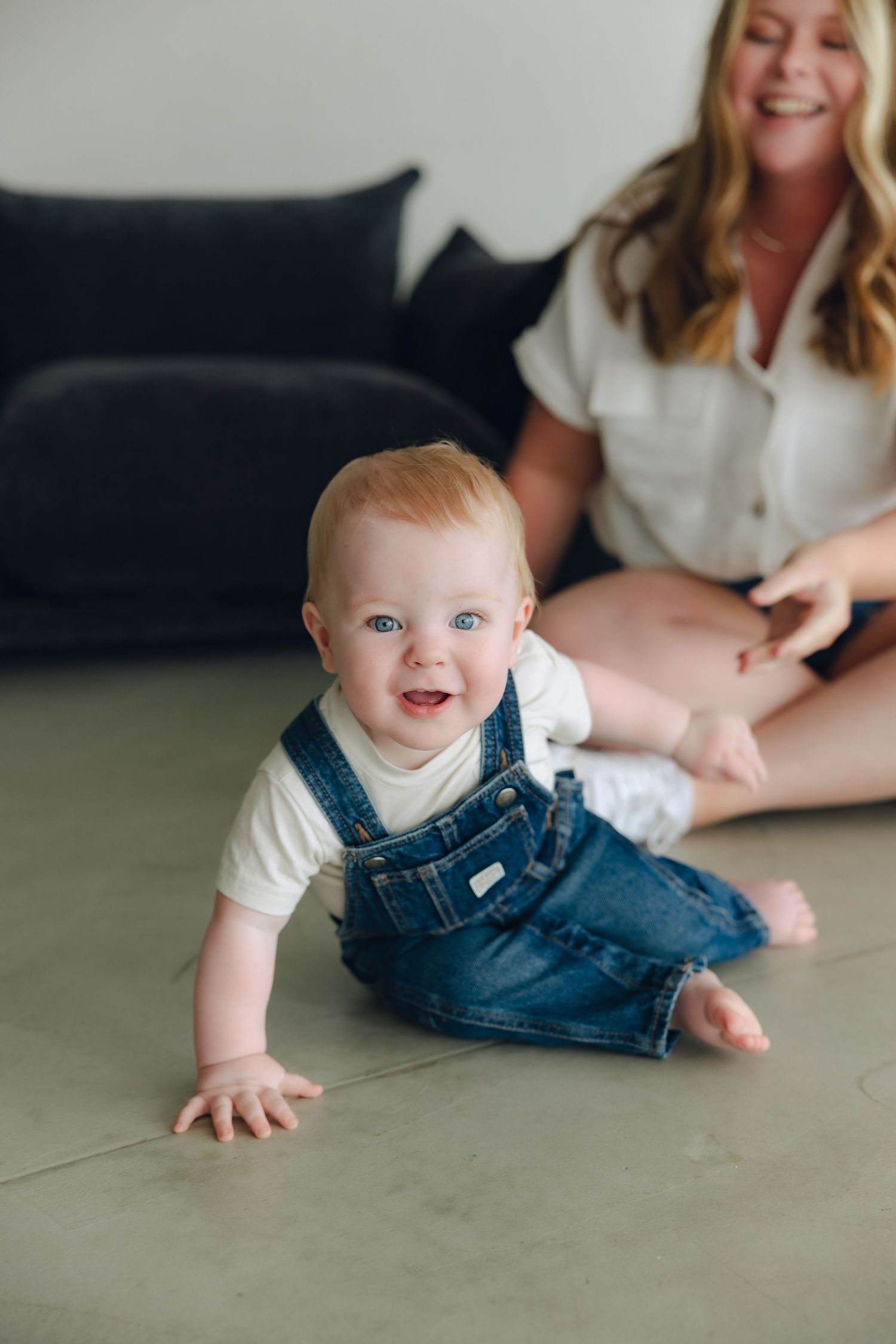 Happy baby in denim overalls crawls on floor while caregiver sits on couch in background.