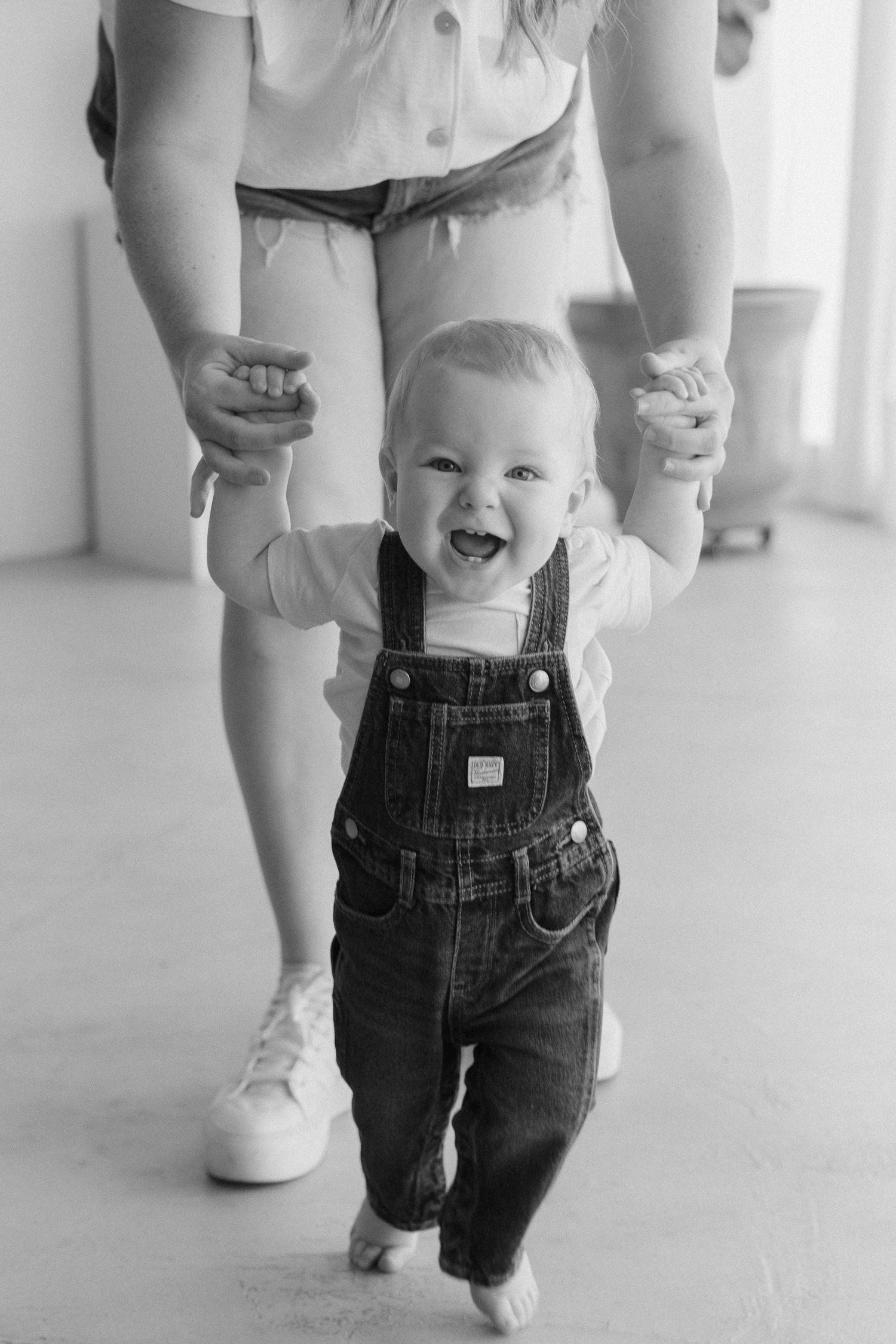 A happy baby in denim overalls takes first steps while being supported by a caregiver in a bright indoor setting.