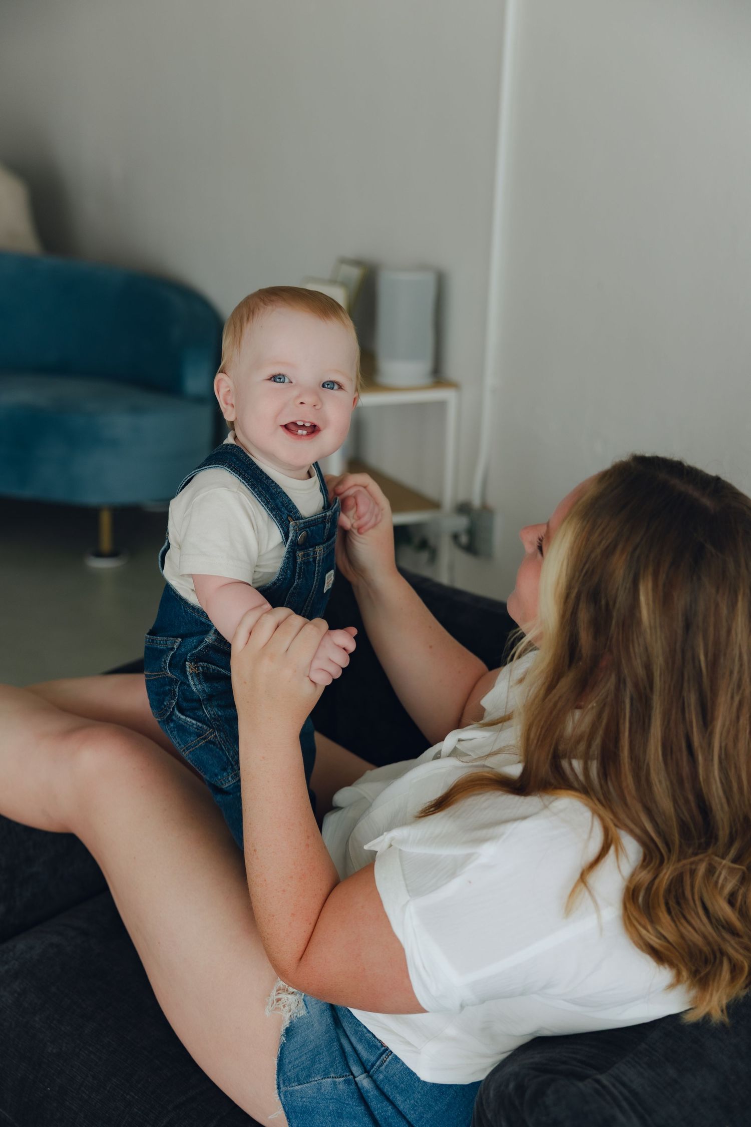An intimate indoor portrait of a parent and baby in overalls sharing a playful moment on a couch.
