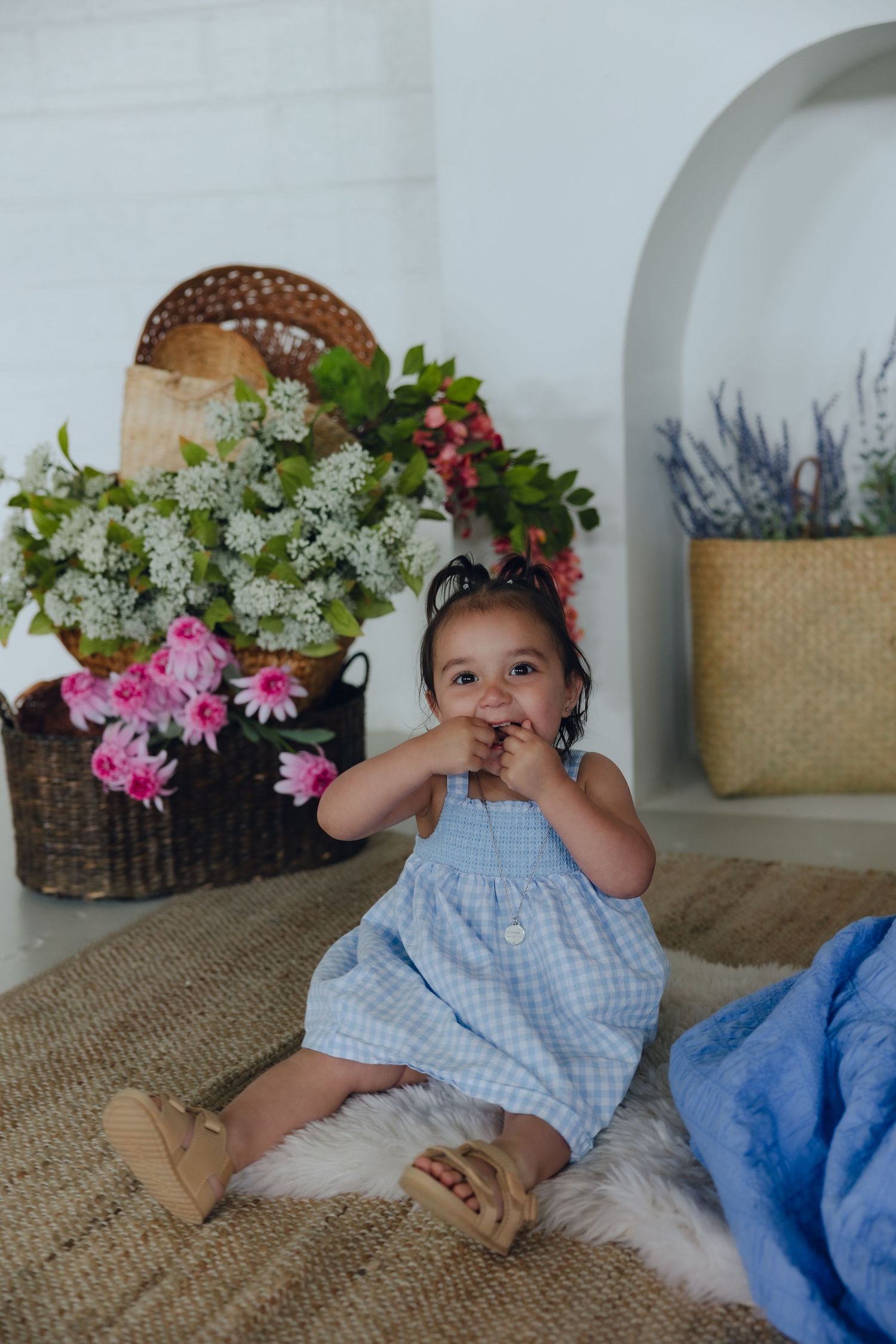 A young child in a blue gingham dress sits on a rug near floral arrangements in white and pink blooms.