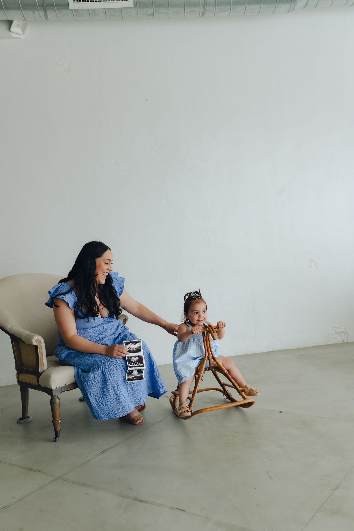 In a minimalist studio space, a woman in denim dress sits beside a child playing on a wooden rocking horse.